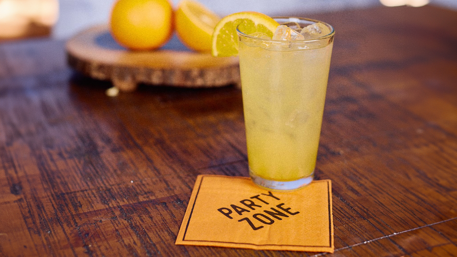 A wooden tabletop featuring a pint glass filled with ice and the Manmosa cocktail. The glass sits upon a party zone napkin and cut irnage on a cutting board are in the background.