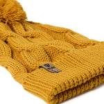 A close up of a  mustard cable-knit beanie hat with a matching pom-pom on top and a ribbed cuff featuring a Till I Die Logo leather tab, shown against a plain white background.