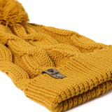 A close up of a  mustard cable-knit beanie hat with a matching pom-pom on top and a ribbed cuff featuring a Till I Die Logo leather tab, shown against a plain white background.