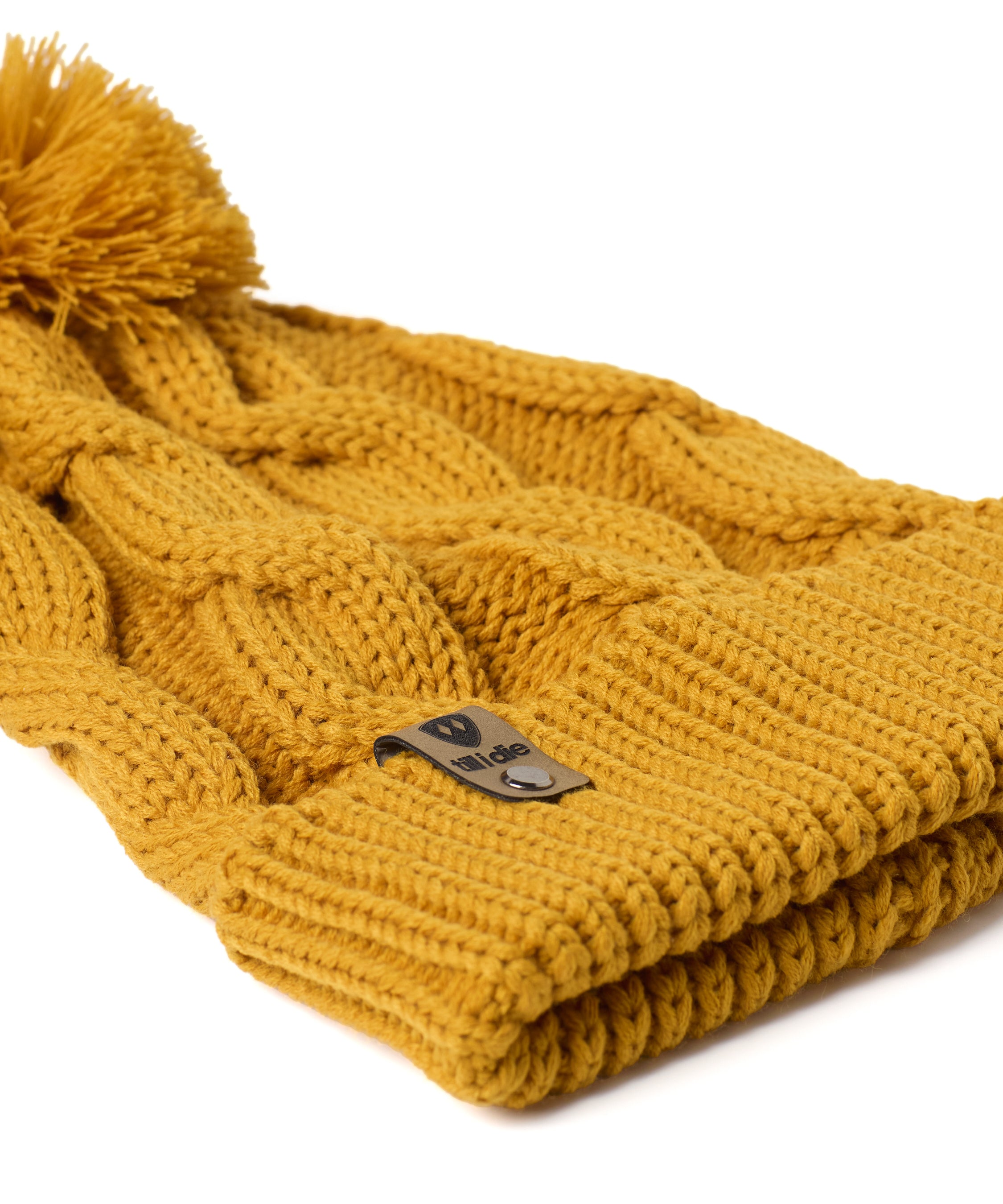 A close up of a  mustard cable-knit beanie hat with a matching pom-pom on top and a ribbed cuff featuring a Till I Die Logo leather tab, shown against a plain white background.