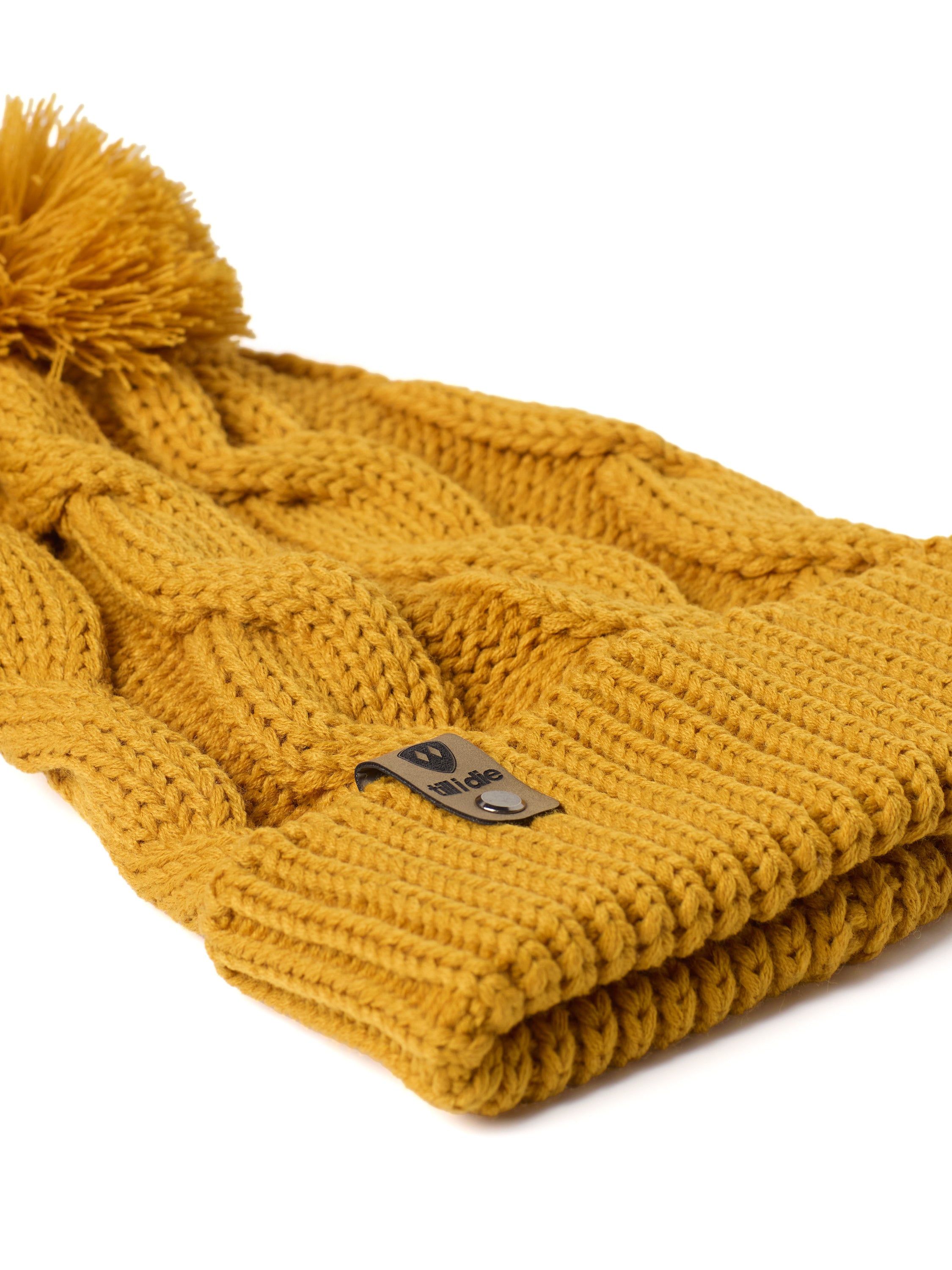 A close up of a  mustard cable-knit beanie hat with a matching pom-pom on top and a ribbed cuff featuring a Till I Die Logo leather tab, shown against a plain white background.