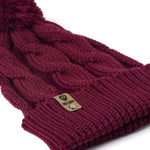 A close up of a burgundy cable-knit beanie hat with a matching pom-pom on top and a ribbed cuff featuring a Till I Die Logo leather tab, shown against a plain white background.