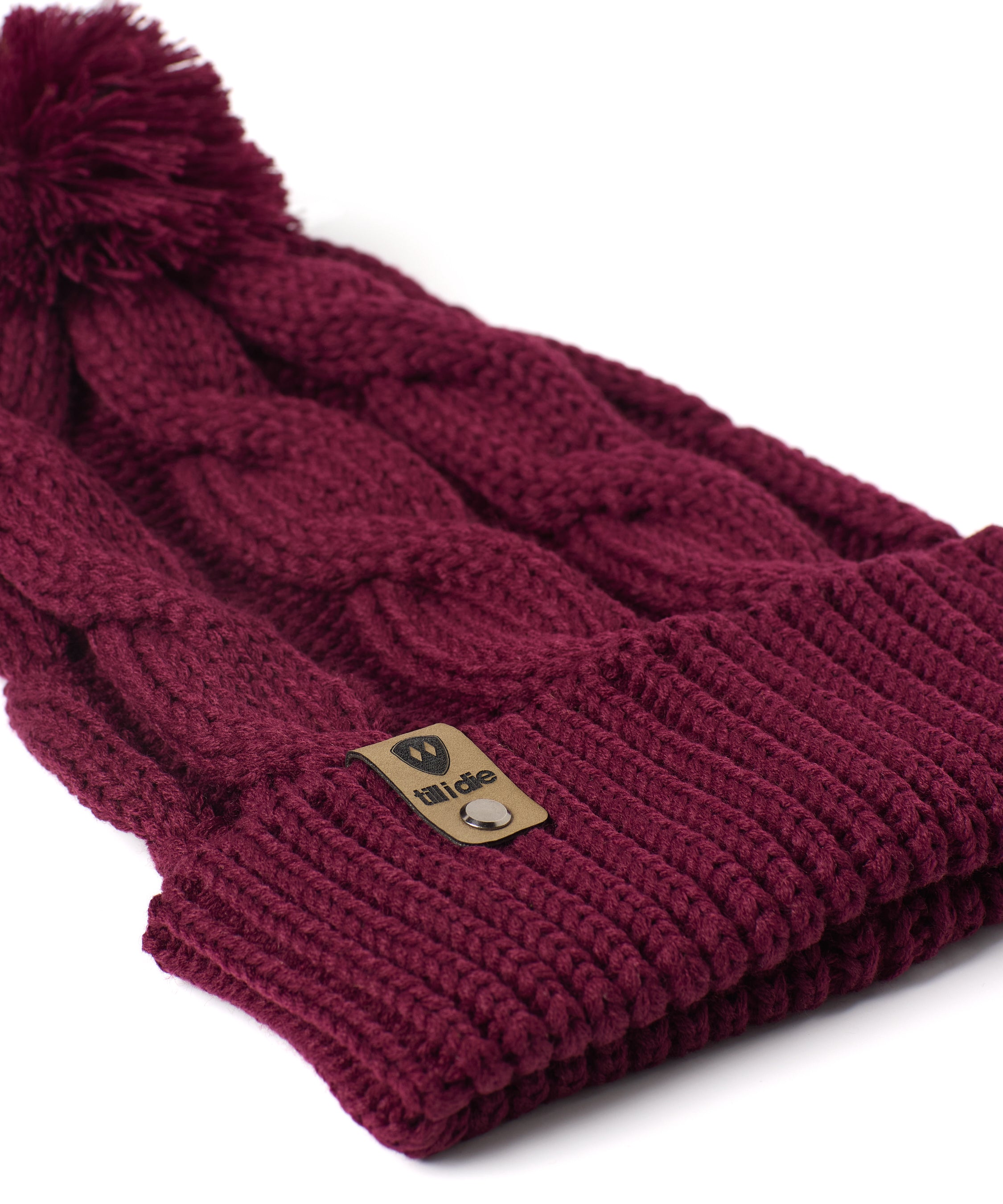 A close up of a burgundy cable-knit beanie hat with a matching pom-pom on top and a ribbed cuff featuring a Till I Die Logo leather tab, shown against a plain white background.