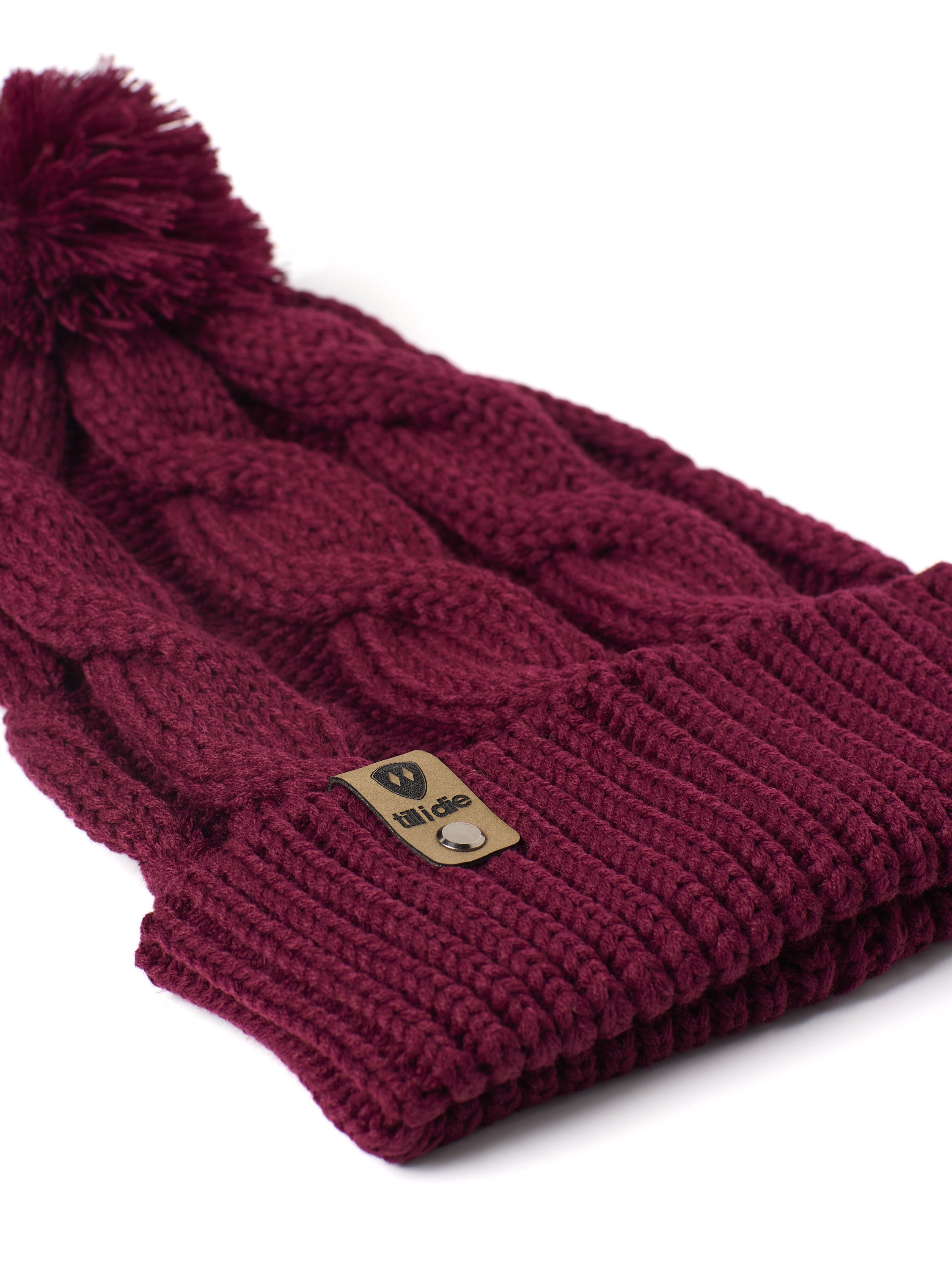 A close up of a burgundy cable-knit beanie hat with a matching pom-pom on top and a ribbed cuff featuring a Till I Die Logo leather tab, shown against a plain white background.