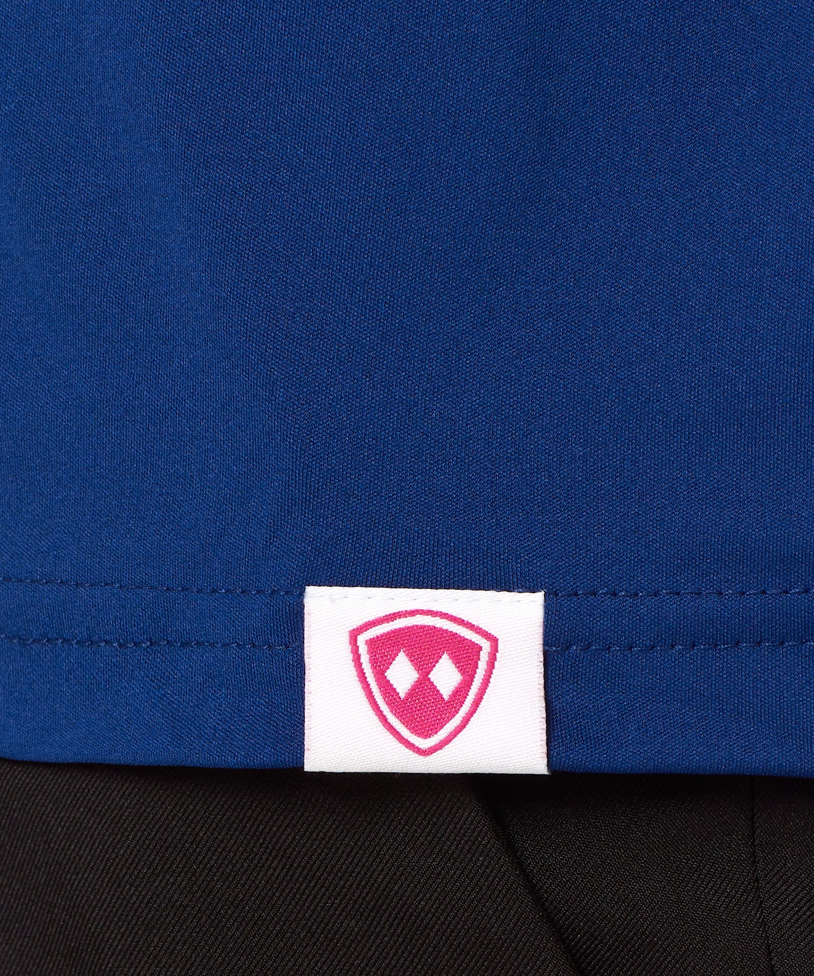 Close-up detail of a pink Double Diamond Shield woven hem label on a blue performance long sleeve shirt.