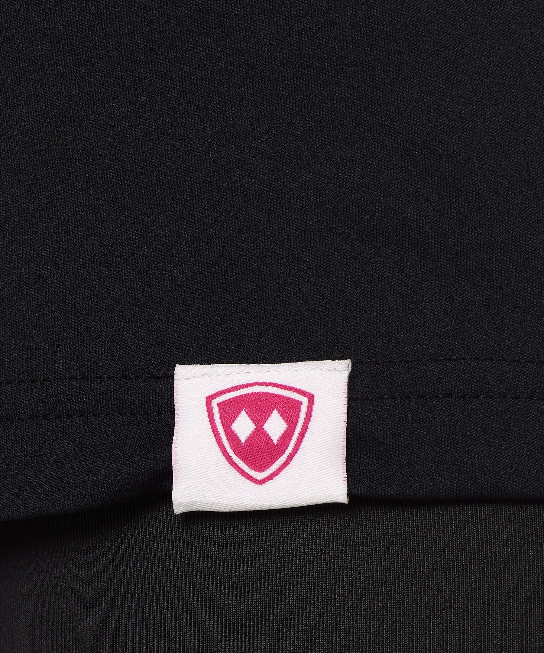 Close-up detail of a pink Double Diamond Shield woven hem label on a black performance long sleeve shirt.