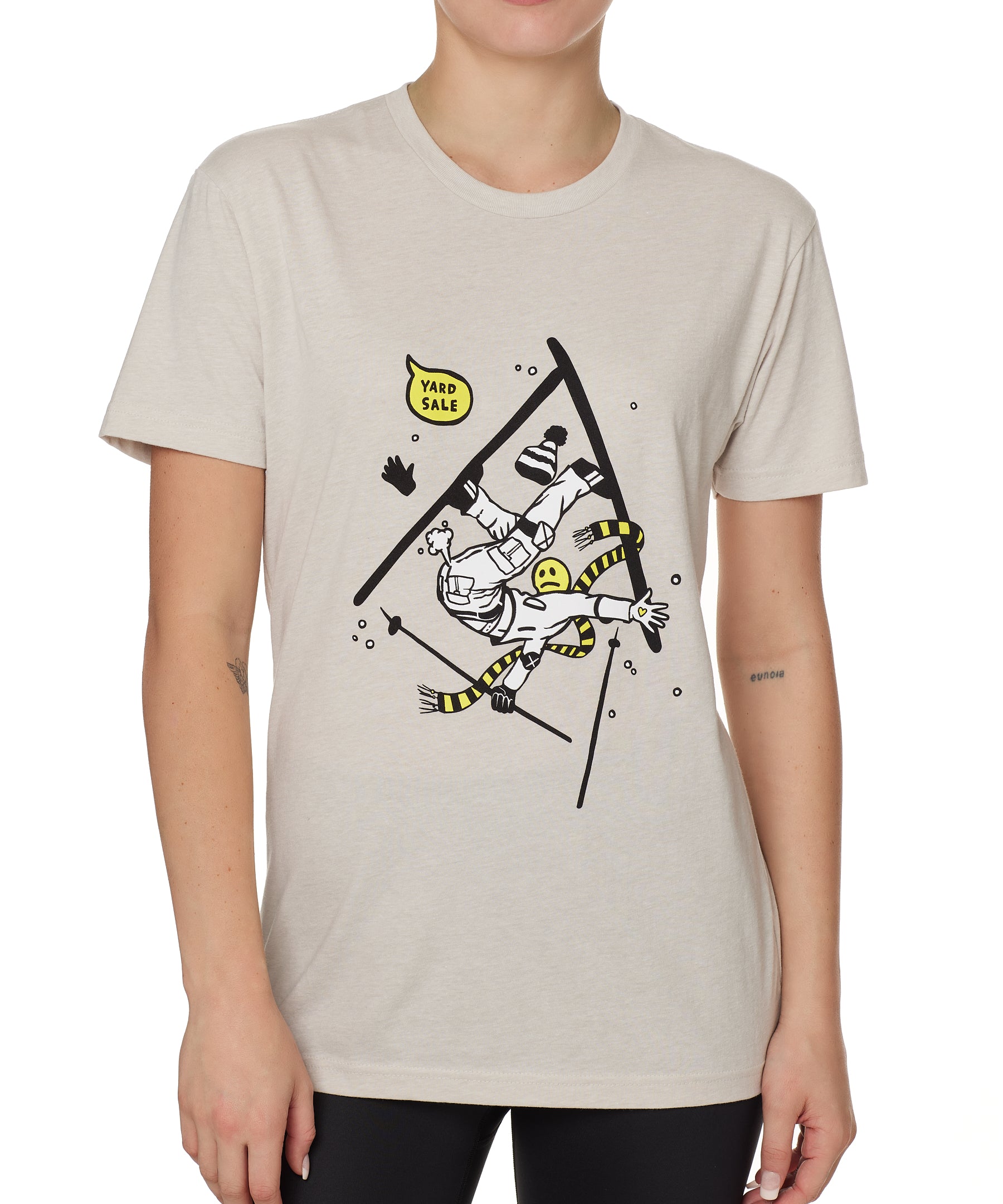 Front view of a female model wearing the Yard Sale crew neck tee in sand, shown on a white background. The graphic features a cartoon skier mid-crash, upside down and losing a hat, gloves, and poles, with a speech bubble that reads “Yard Sale.”