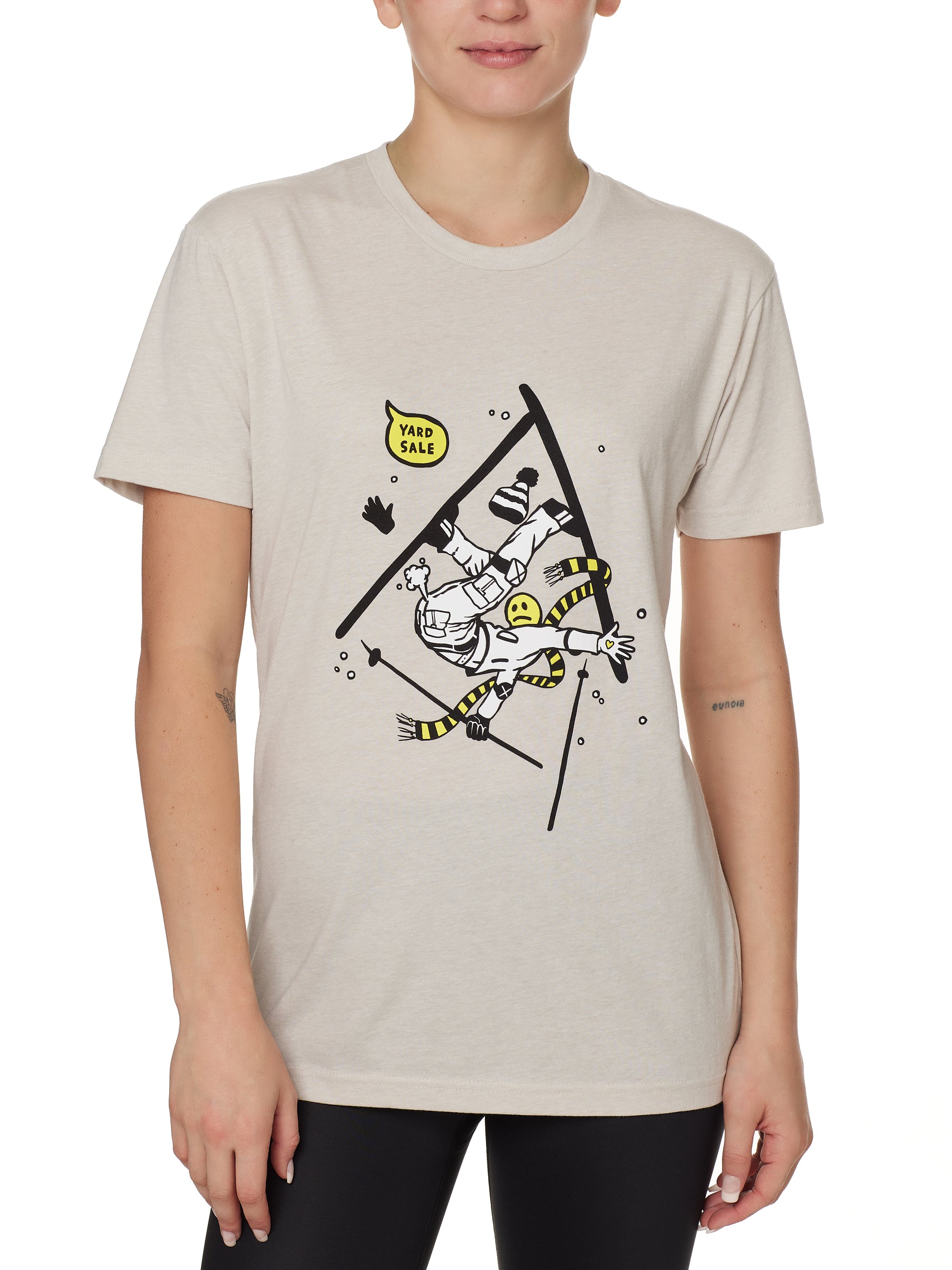 Front view of a female model wearing the Yard Sale crew neck tee in sand, shown on a white background. The graphic features a cartoon skier mid-crash, upside down and losing a hat, gloves, and poles, with a speech bubble that reads “Yard Sale.”