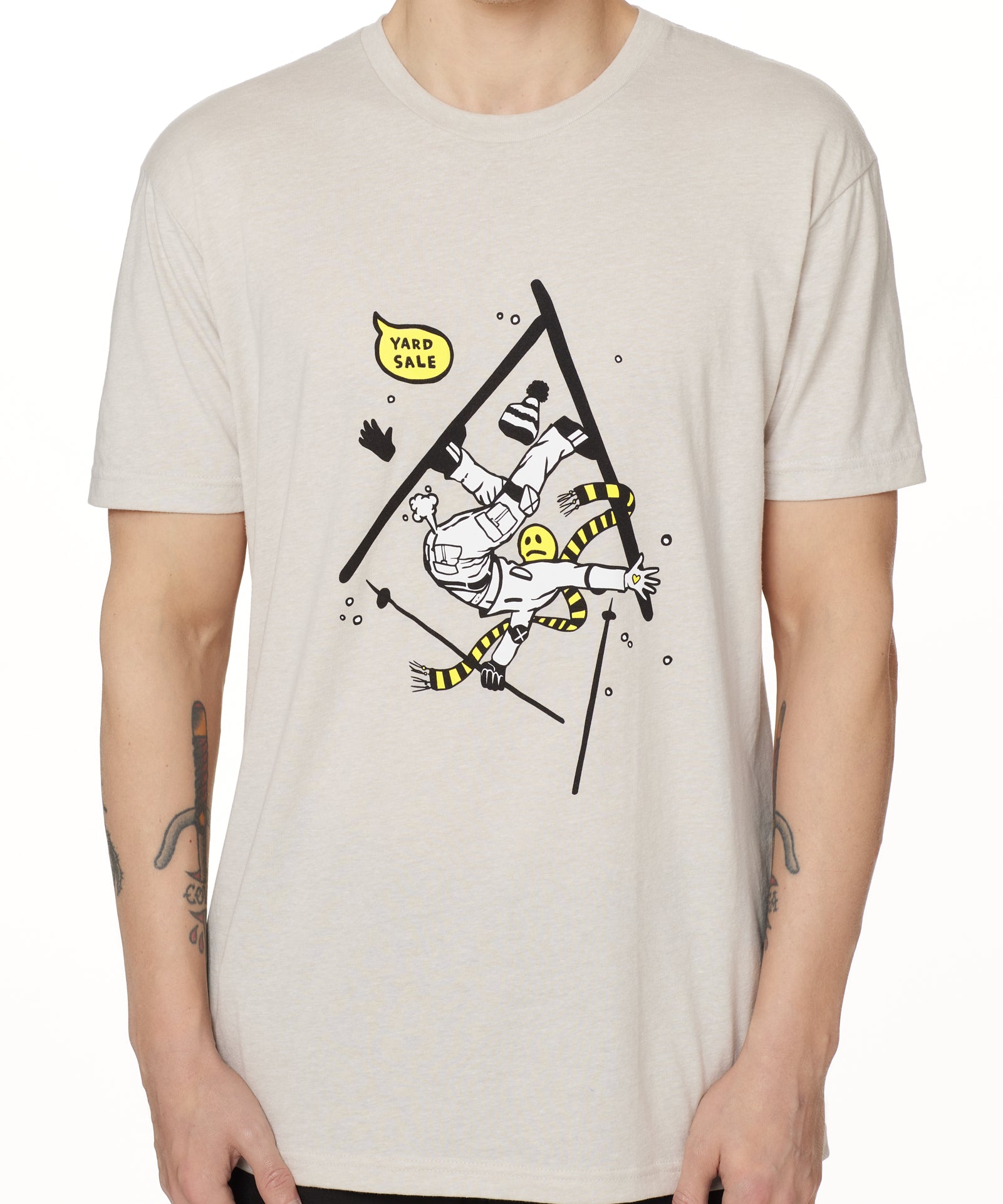 Front view of a male model wearing the Yard Sale crew neck tee in sand, shown on a white background. The graphic features a cartoon skier mid-crash, upside down and losing a hat, gloves, and poles, with a speech bubble that reads “Yard Sale.”