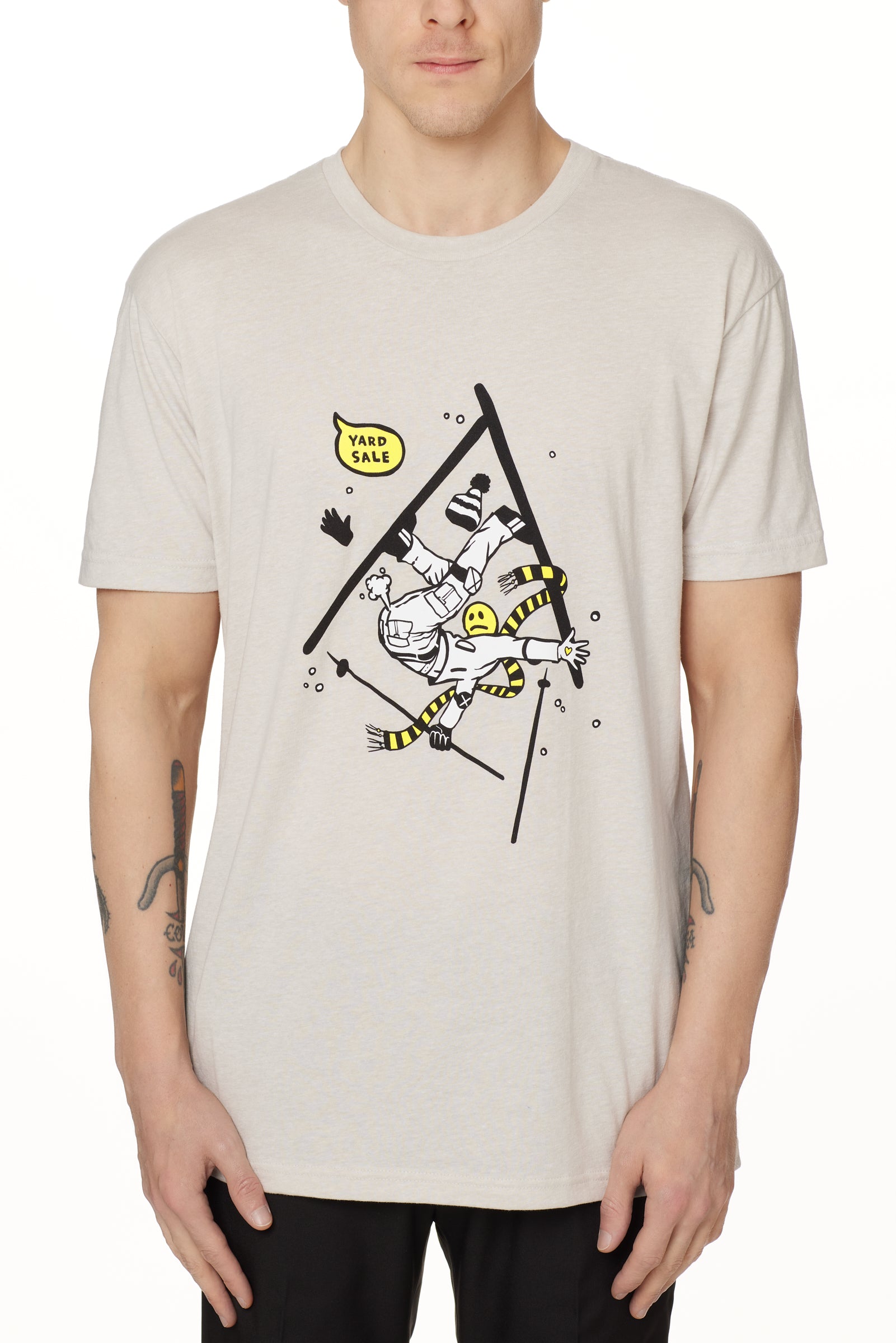 Front view of a male model wearing the Yard Sale crew neck tee in sand, shown on a white background. The graphic features a cartoon skier mid-crash, upside down and losing a hat, gloves, and poles, with a speech bubble that reads “Yard Sale.”
