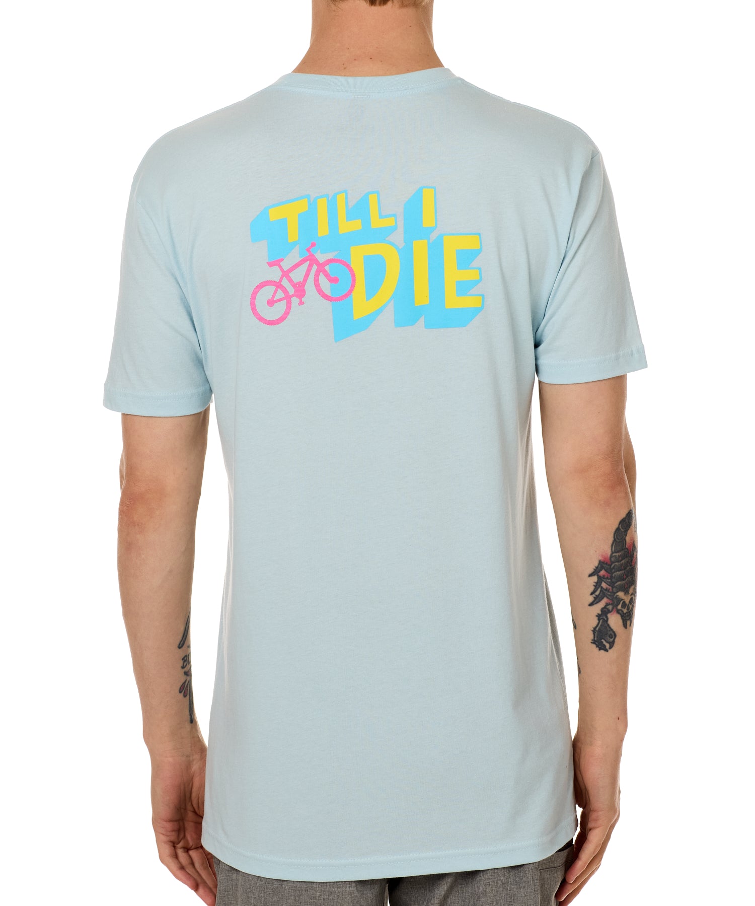 Back view of a male model wearing the Bike till i die tee shirt in light blue on a white background. The graphic features a pink mountain bike layered over bold blue and yellow till i die text