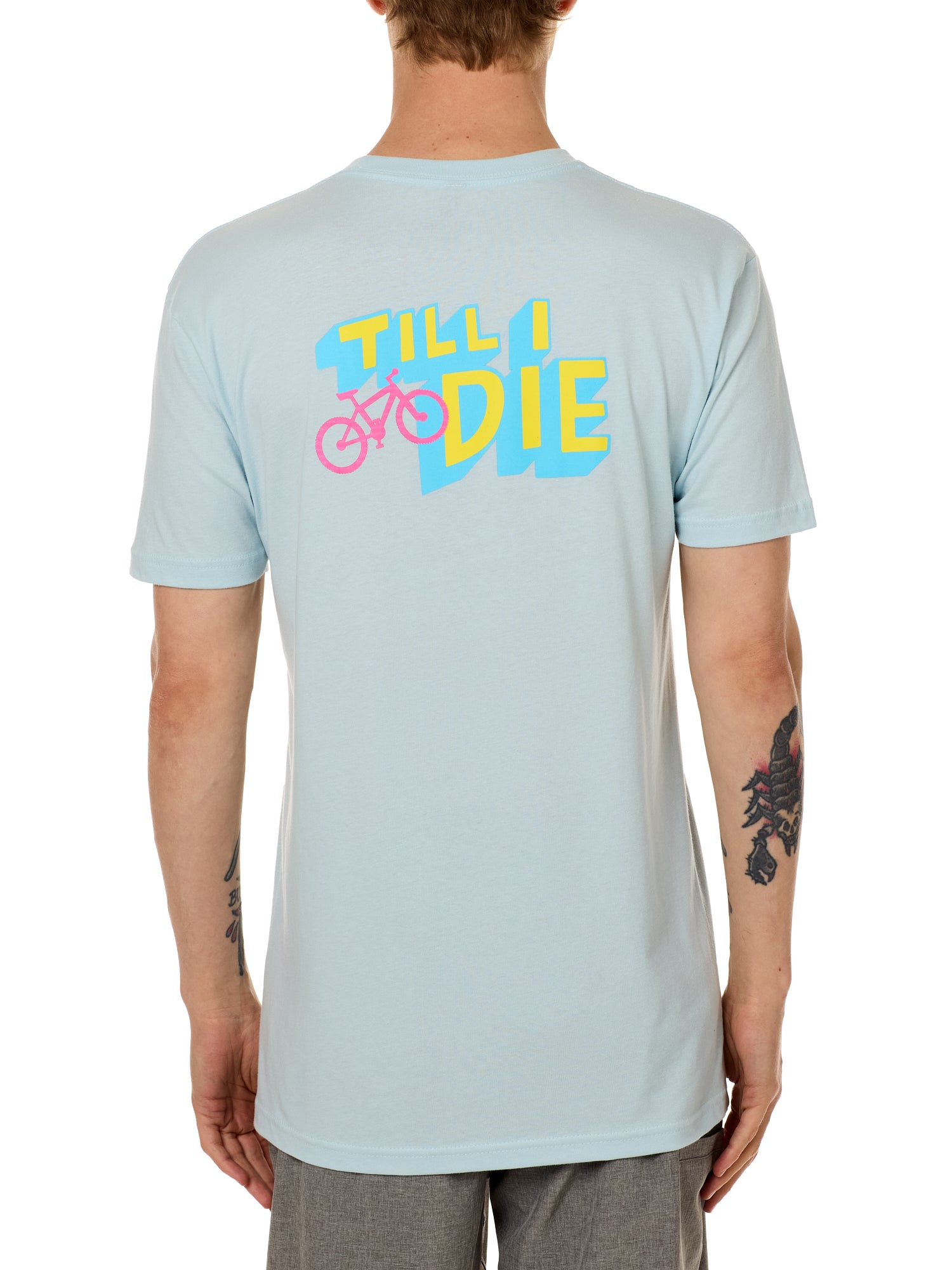 Back view of a male model wearing the Bike till i die tee shirt in light blue on a white background. The graphic features a pink mountain bike layered over bold blue and yellow till i die text