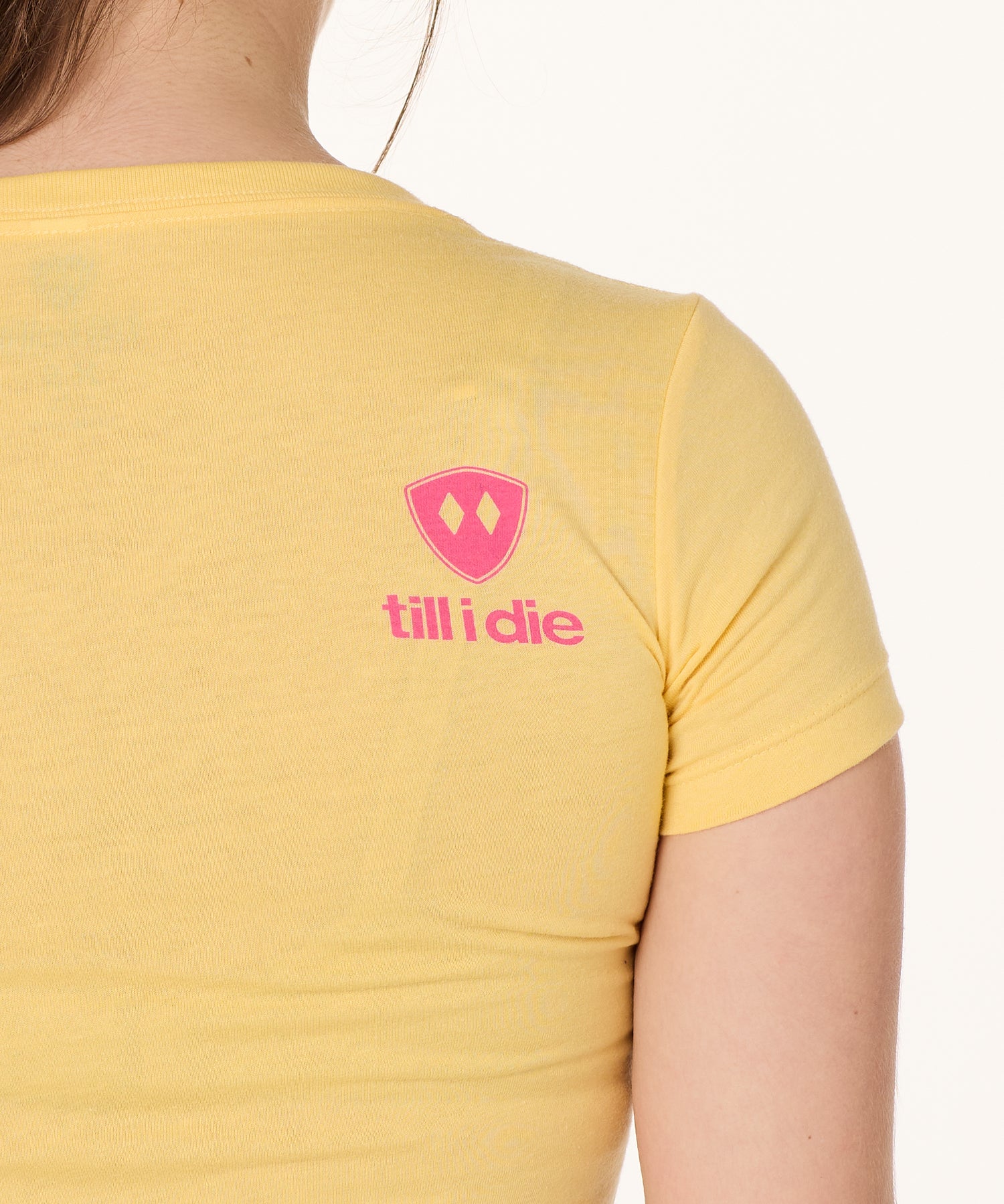 Back view of a female model wearing a Banana short sleeve shirt on a white background, featuring a hot pink Till I Die logo on the wearer’s top right shoulder.