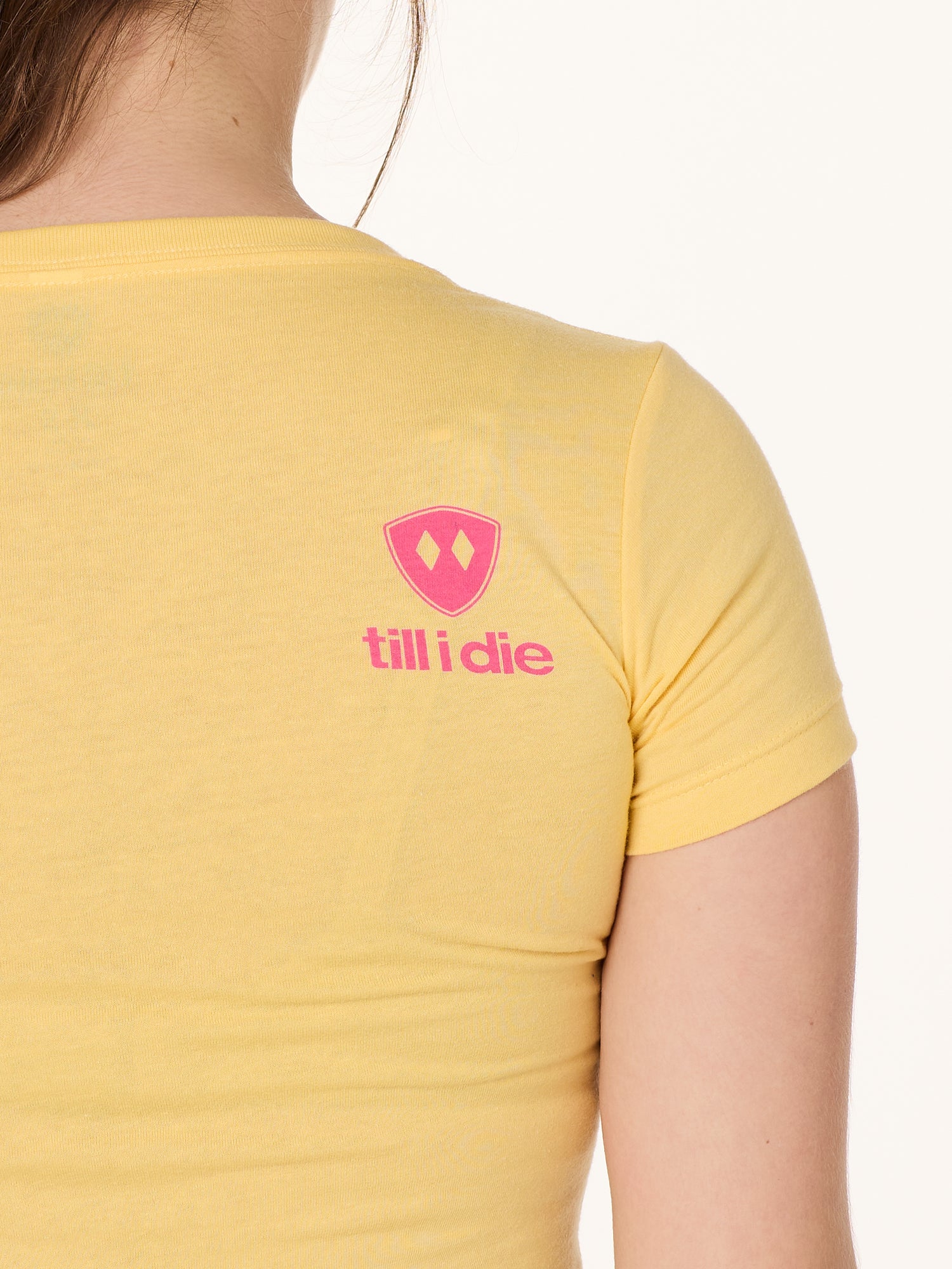 Back view of a female model wearing a Banana short sleeve shirt on a white background, featuring a hot pink Till I Die logo on the wearer’s top right shoulder.