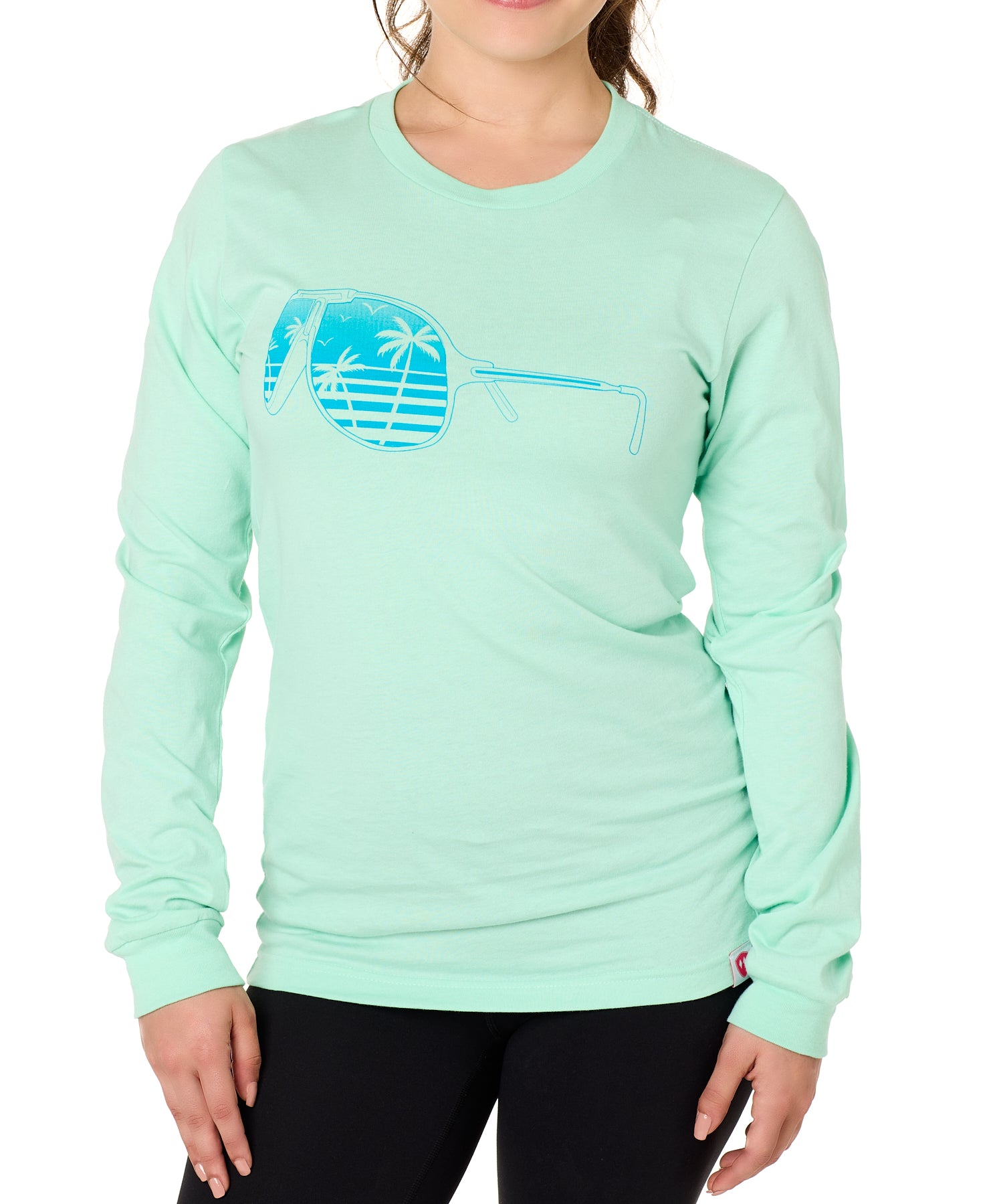 Front view of a female model wearing a long sleeve crew neck Mint Green T-Shirt. The graphic features a blue print of sunglasses with a tropical beach scene reflected in the lenses.