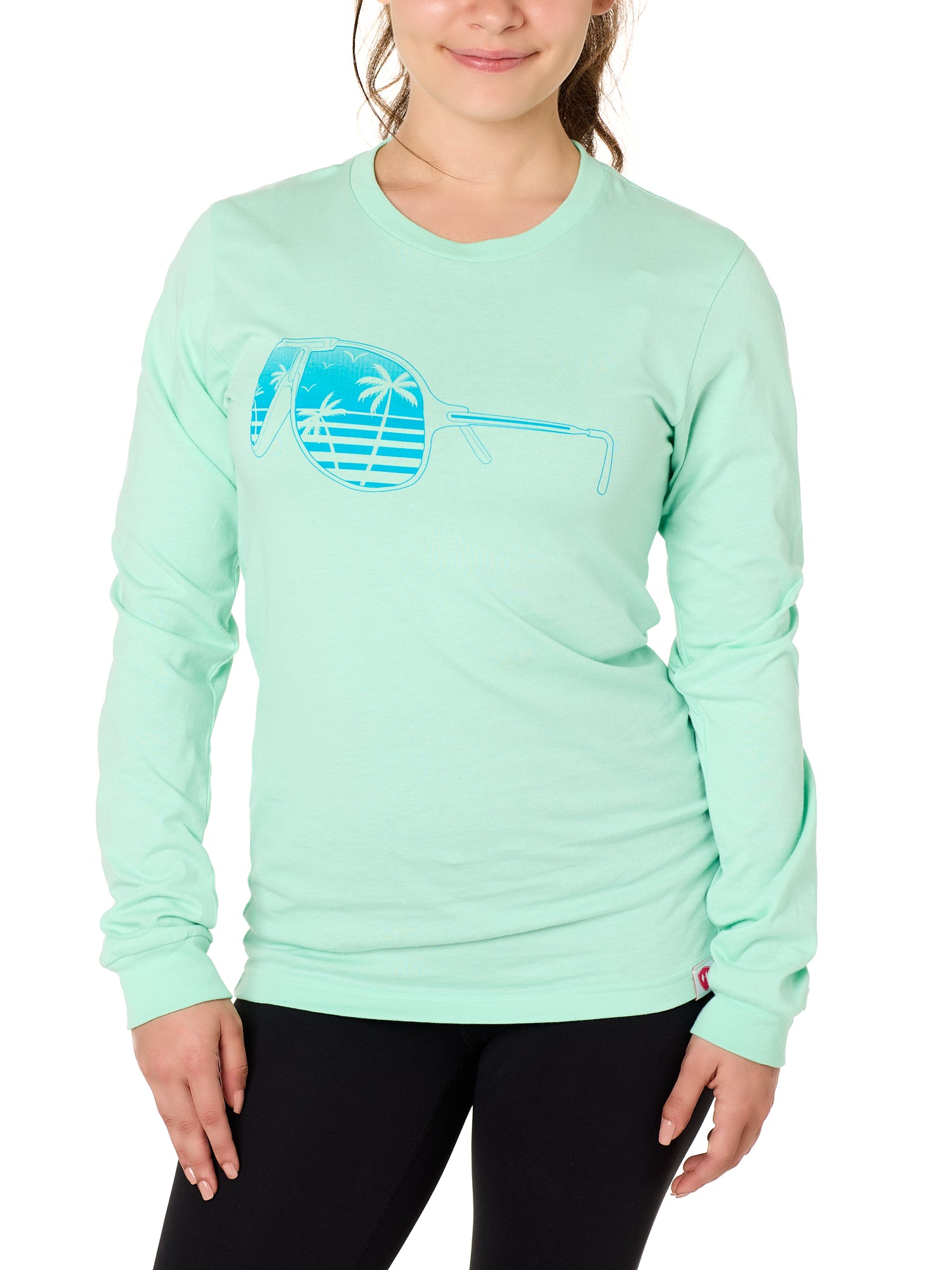 Front view of a female model wearing a long sleeve crew neck Mint Green T-Shirt. The graphic features a blue print of sunglasses with a tropical beach scene reflected in the lenses.