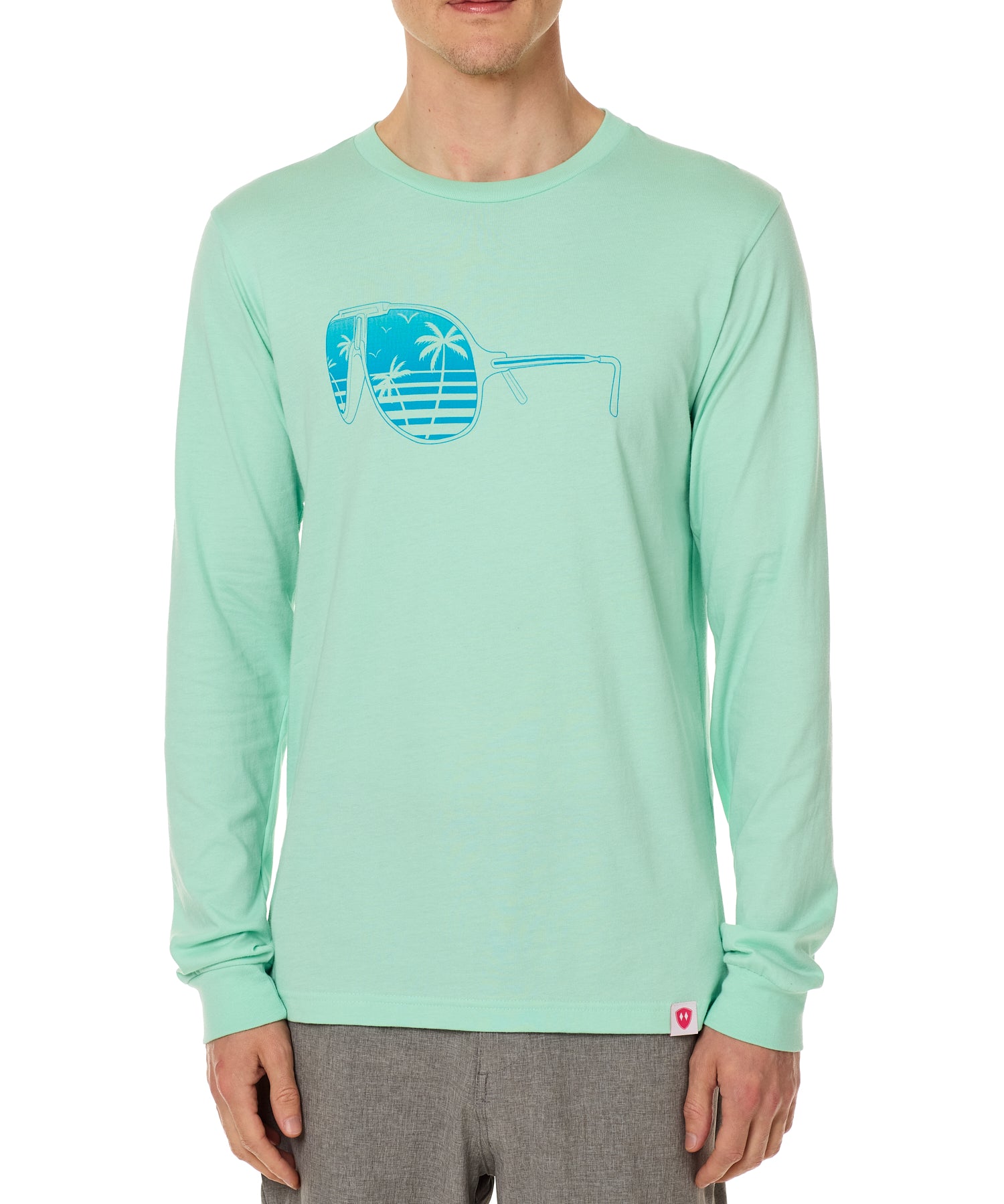 Front view of a male model wearing a long sleeve crew neck Mint Green T-Shirt. The graphic features a blue print of sunglasses with a tropical beach scene reflected in the lenses.