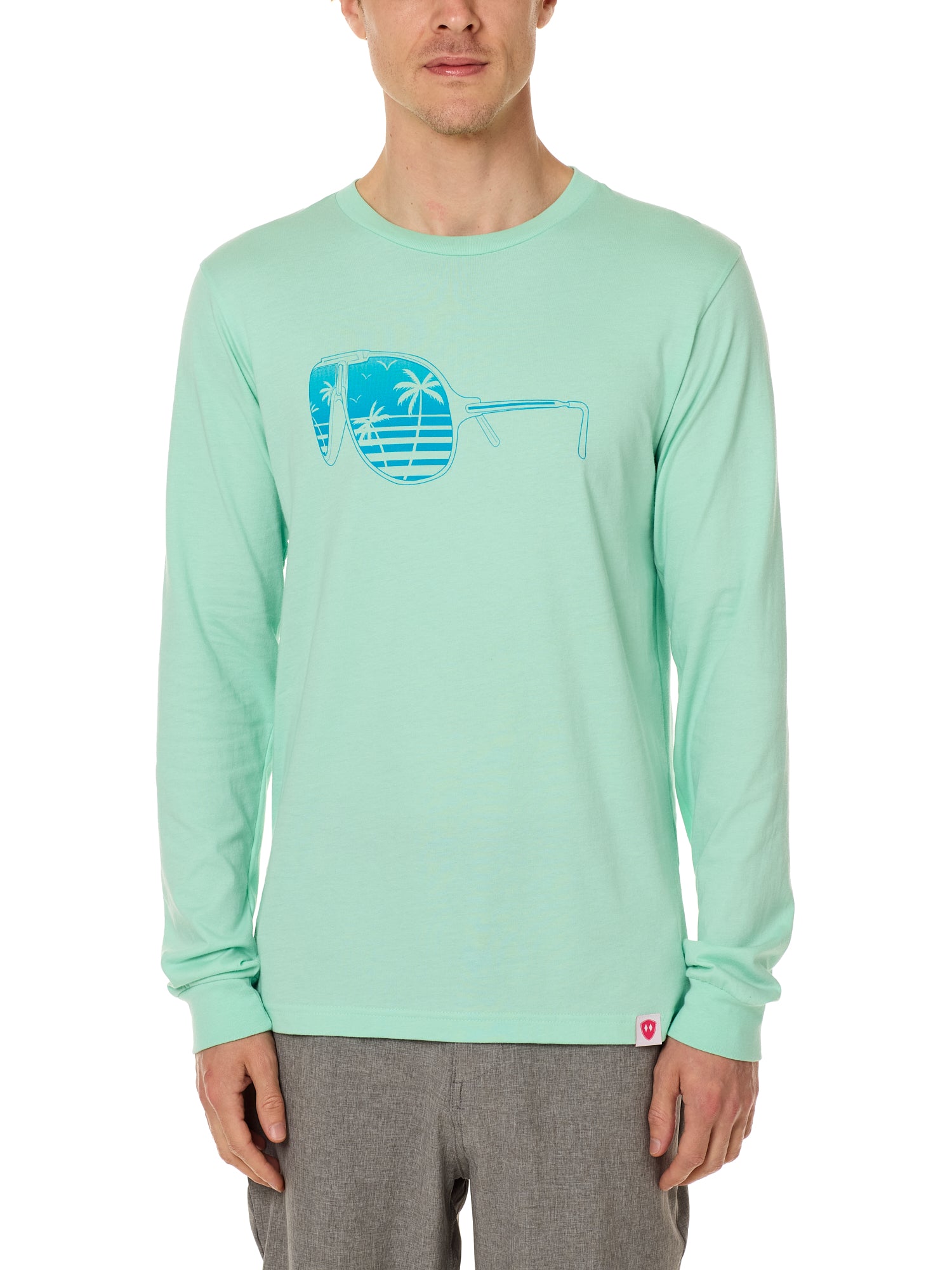 Front view of a male model wearing a long sleeve crew neck Mint Green T-Shirt. The graphic features a blue print of sunglasses with a tropical beach scene reflected in the lenses.