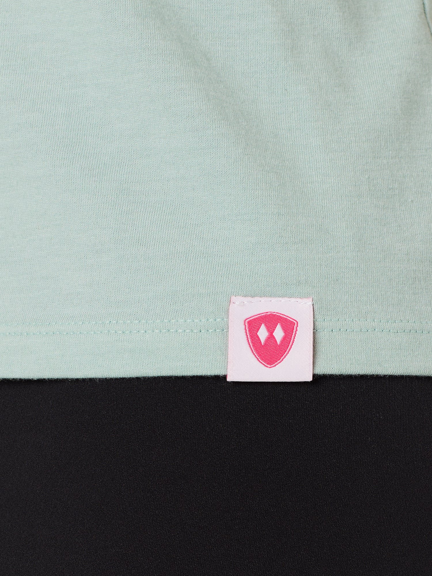 Close-up detail of a pink Double Diamond Shield woven hem label on a dusty blue tank.