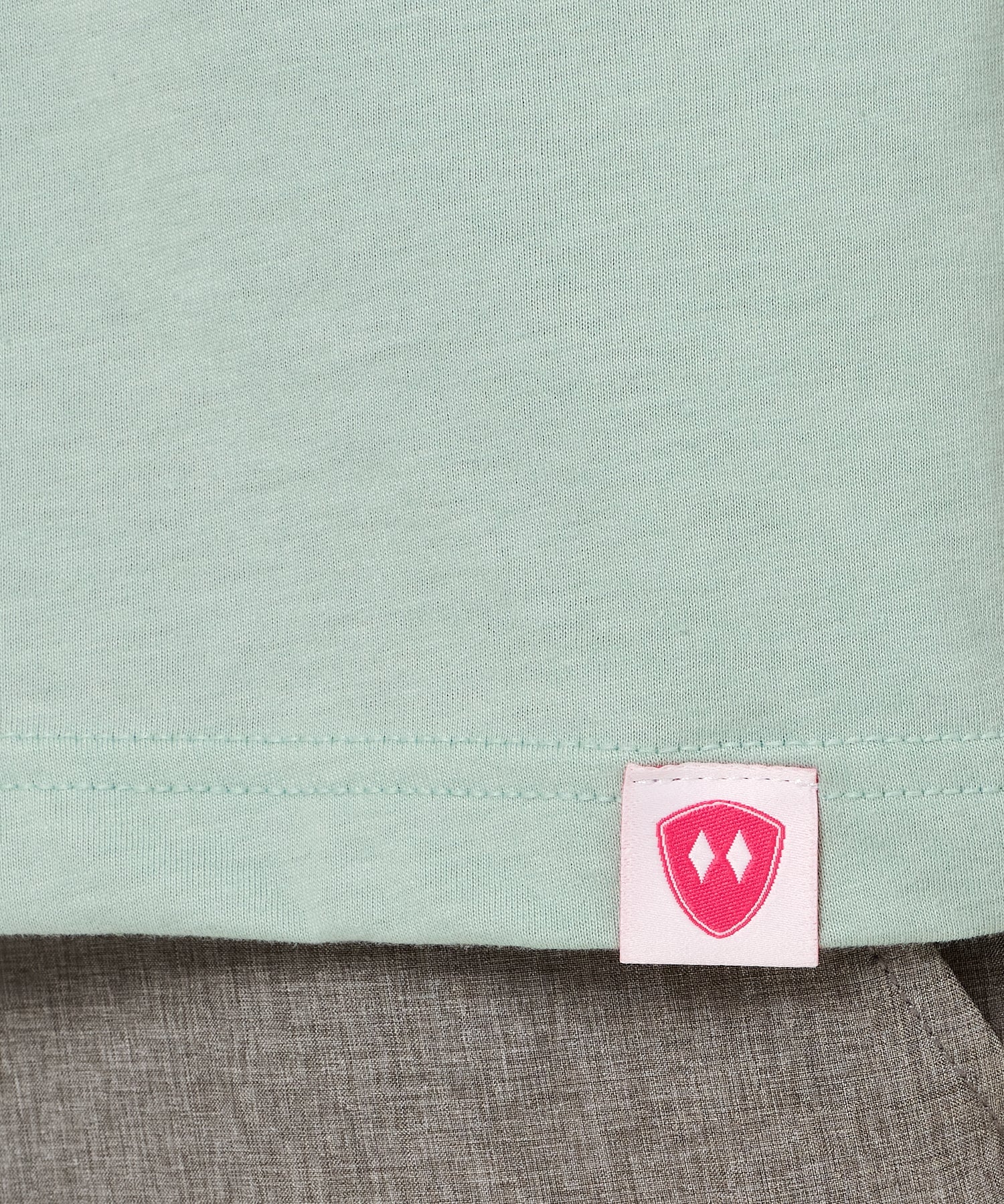 Close-up detail of a pink Double Diamond Shield woven hem label on a dusty blue crew neck tee.