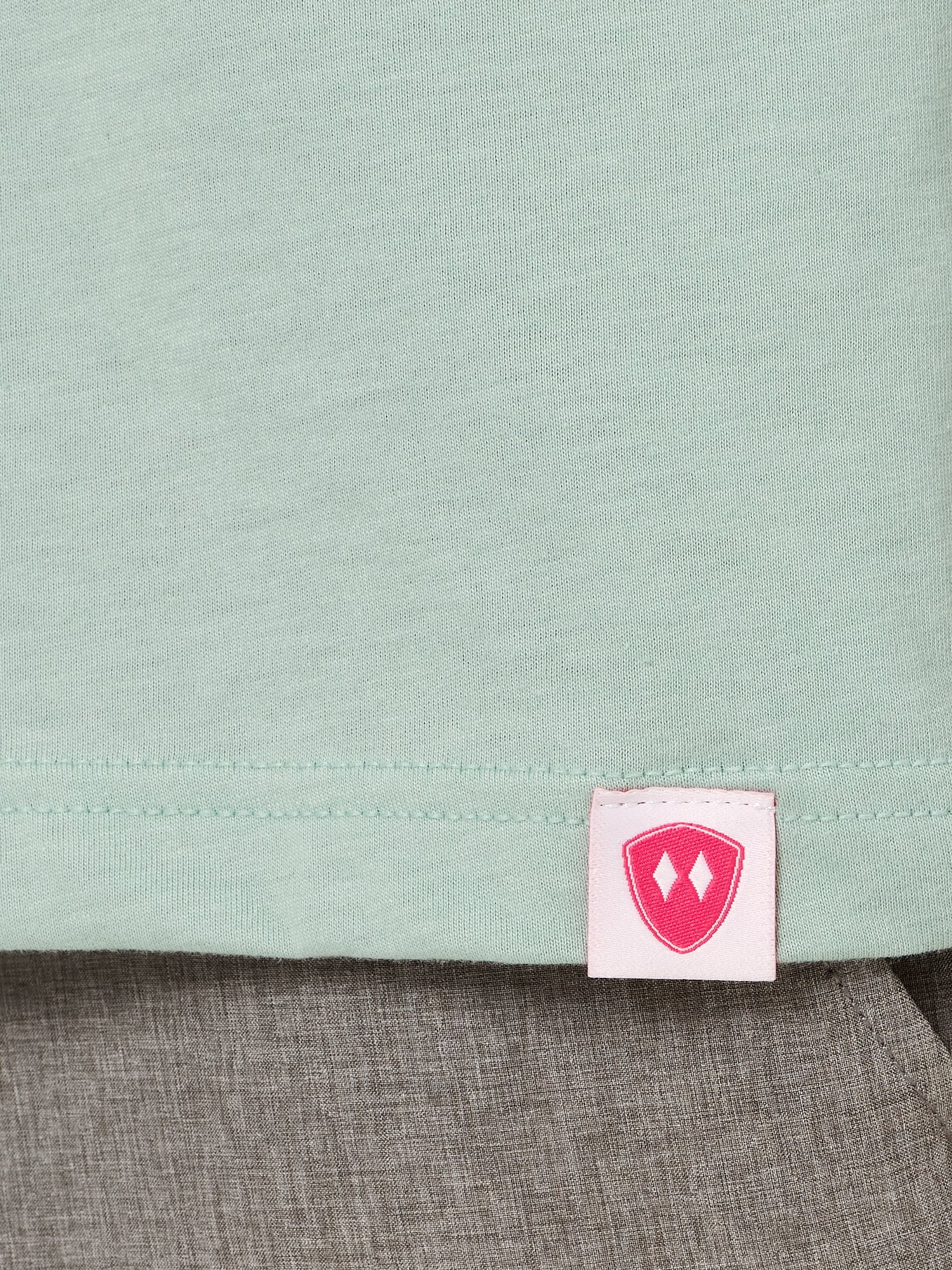 Close-up detail of a pink Double Diamond Shield woven hem label on a dusty blue crew neck tee.