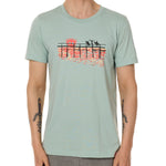 Front view of a male model wearing the Endless Summer crew neck tee in dusty blue, displayed on a white background. The graphic features three silhouetted surfers walking along an ocean boardwalk, with a setting sun—stylized as the Till I Die double diamond shield—glowing in the background.