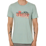 Front view of a male model wearing the Endless Summer crew neck tee in dusty blue, displayed on a white background. The graphic features three silhouetted surfers walking along an ocean boardwalk, with a setting sun—stylized as the Till I Die double diamond shield—glowing in the background.