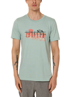 Front view of a male model wearing the Endless Summer crew neck tee in dusty blue, displayed on a white background. The graphic features three silhouetted surfers walking along an ocean boardwalk, with a setting sun—stylized as the Till I Die double diamond shield—glowing in the background.