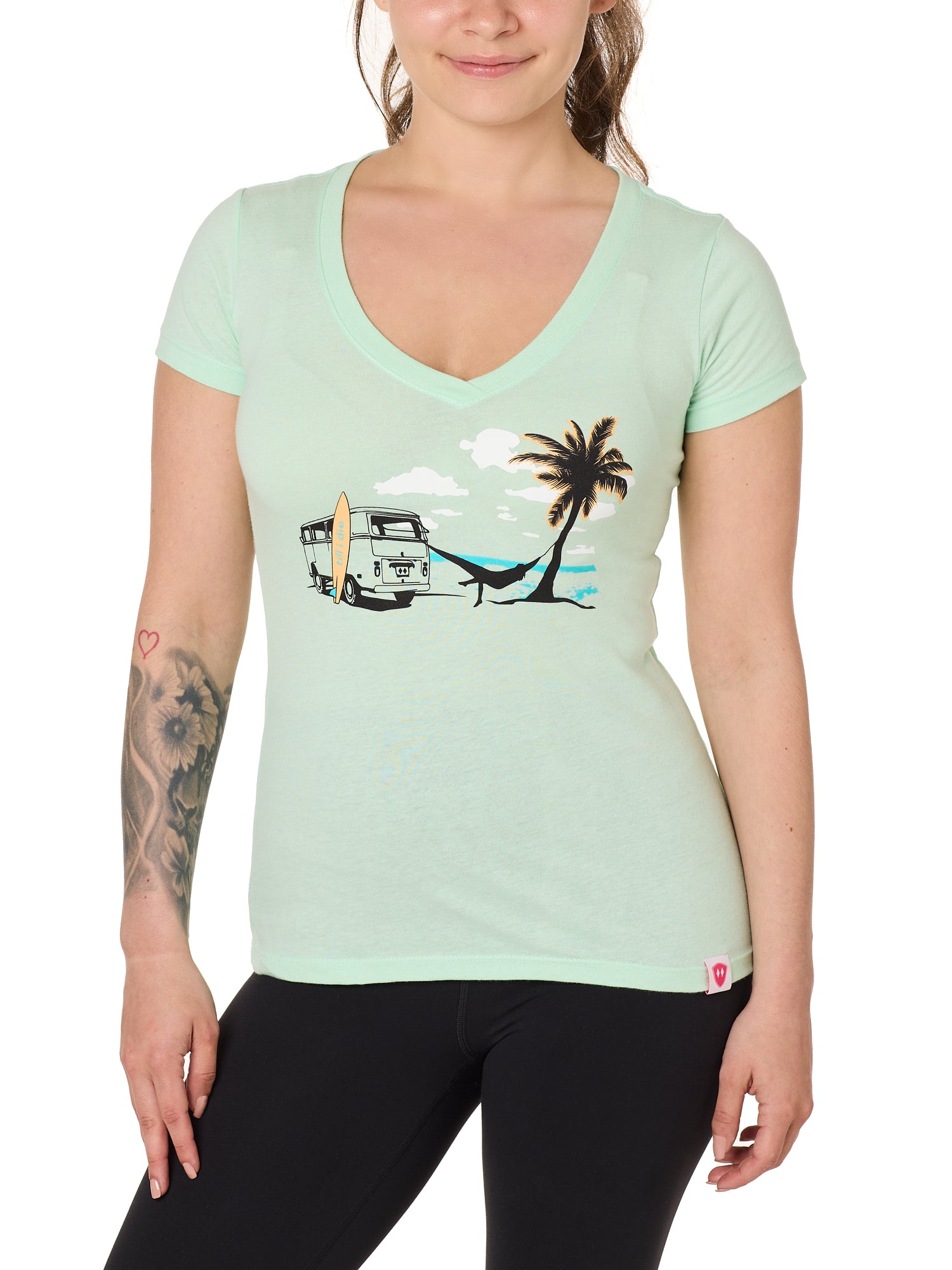 Front view of a female model wearing the Hammock V-neck tee in mint, displayed on a white background. The graphic depicts a vintage VW bus parked on a beach, with a surfboard leaning against it and a silhouetted figure relaxing in a hammock strung between the van and a palm tree.