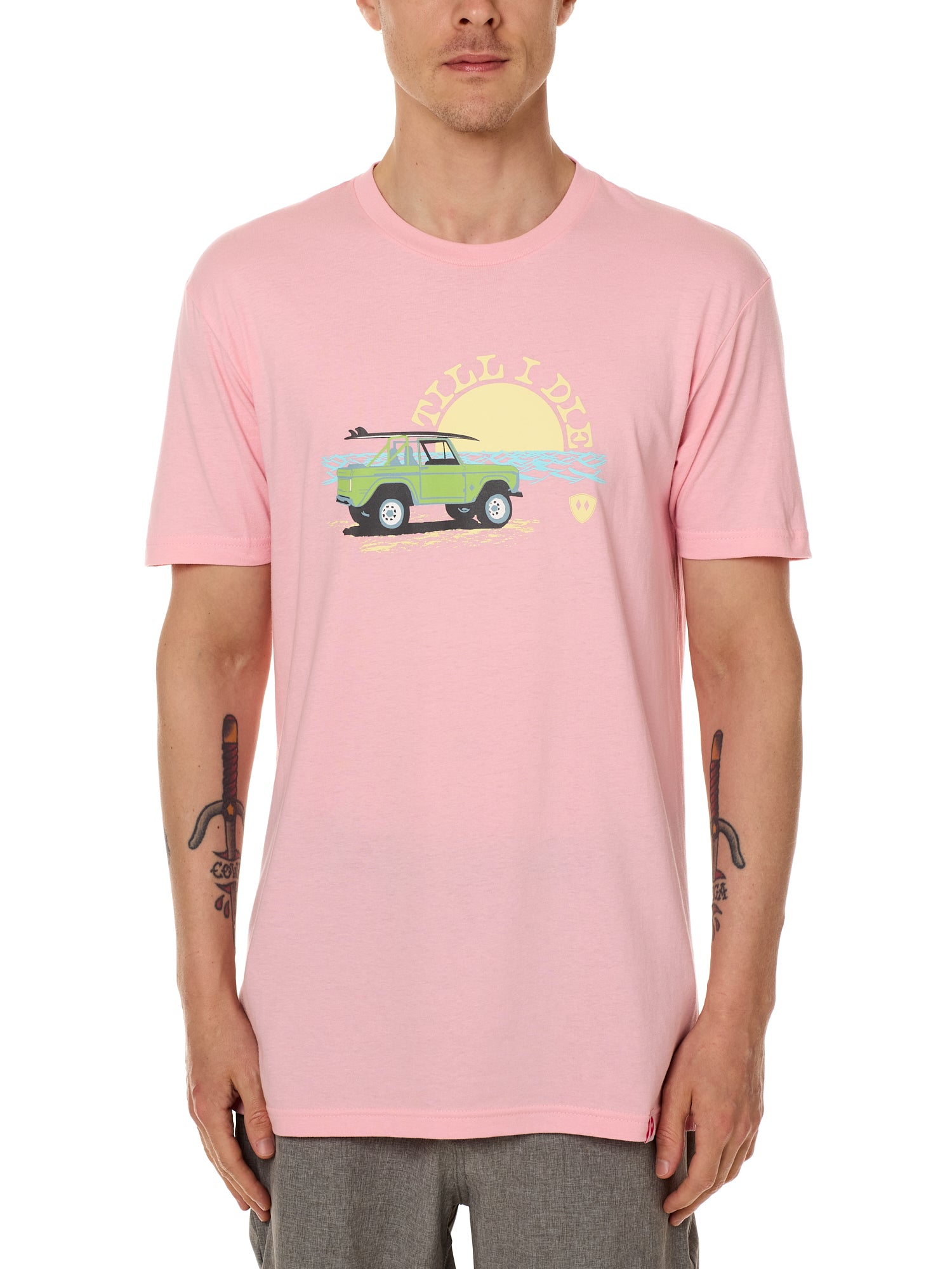 Front view of a male model wearing the Surf Truck crew neck tee in light pink on a white background. The graphic features a classic green Bronco with a surfboard on the roof, parked in front of an ocean sunset, arched by “Till I Die” text.