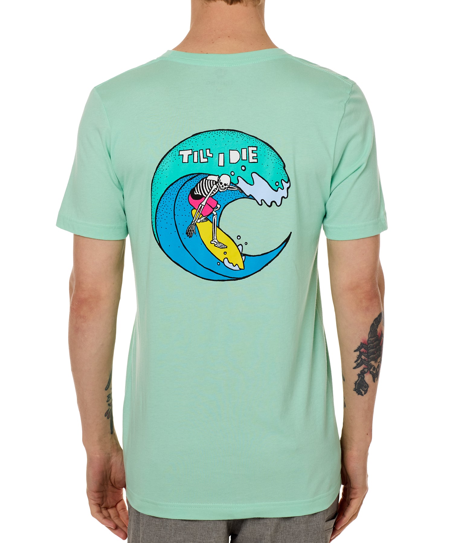 Back view of a male model wearing the Surfer Skeleton tee in mint green on a white background. The graphic features a multicolored circular graphic of a skeleton surfing a wave, with “Till I Die” text at the top.