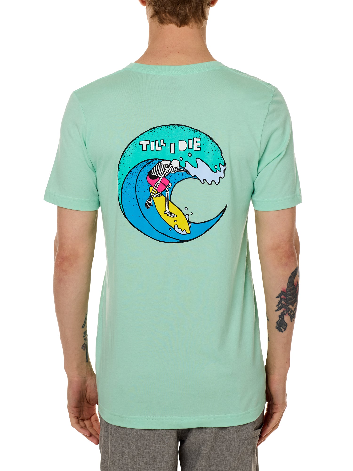 Back view of a male model wearing the Surfer Skeleton tee in mint green on a white background. The graphic features a multicolored circular graphic of a skeleton surfing a wave, with “Till I Die” text at the top.