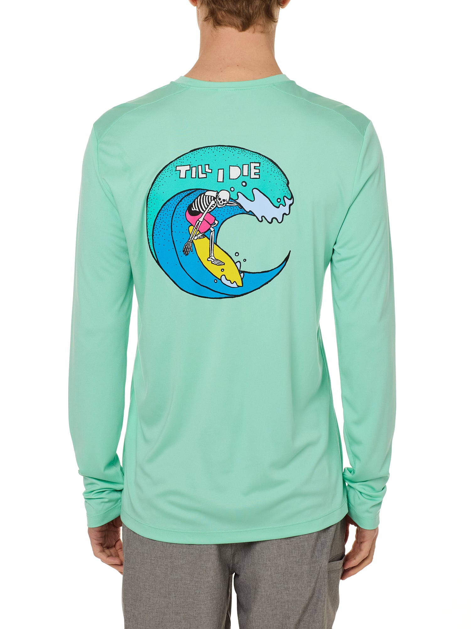 Back view of a male model wearing the Surfer Skeleton performance long sleeve tee in mint green on a white background. The graphic features a multicolored circular graphic of a skeleton surfing a wave, with “Till I Die” text at the top.