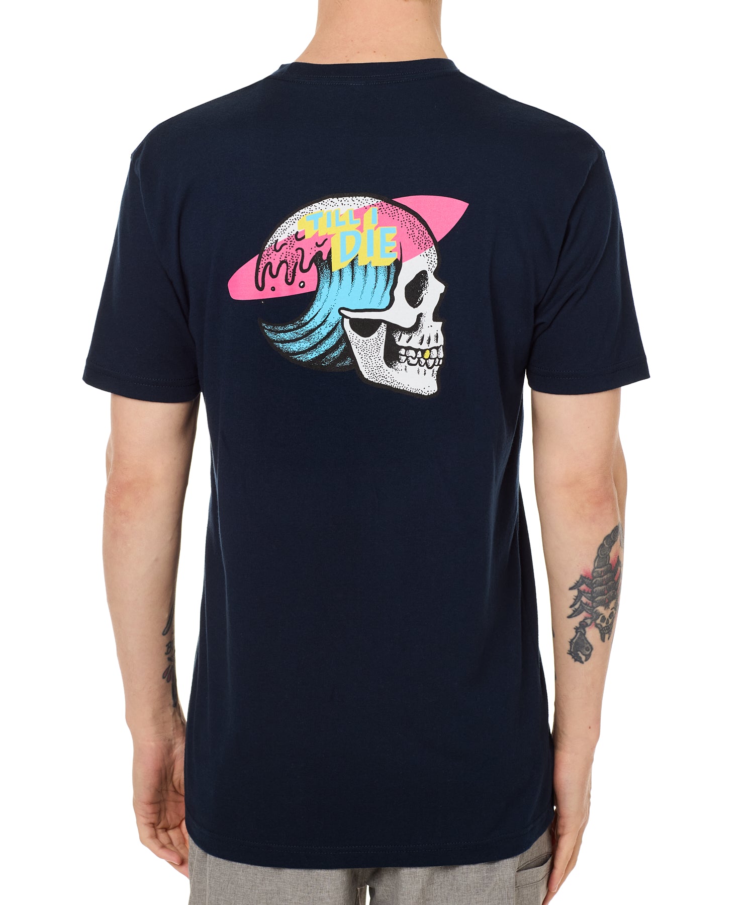 Back view of a male model wearing the Skull Wave Head Tee in navy. The back graphic features a skull with a wave inside the head, overlapped by a pink surfboard and accented with yellow and blue "Till I Die" text.