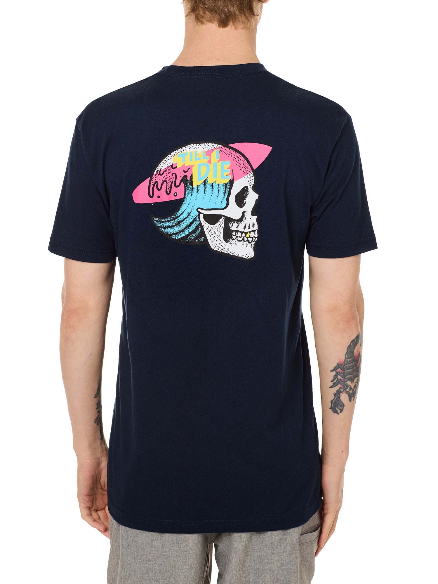 Back view of a male model wearing the Skull Wave Head Tee in navy. The back graphic features a skull with a wave inside the head, overlapped by a pink surfboard and accented with yellow and blue "Till I Die" text.