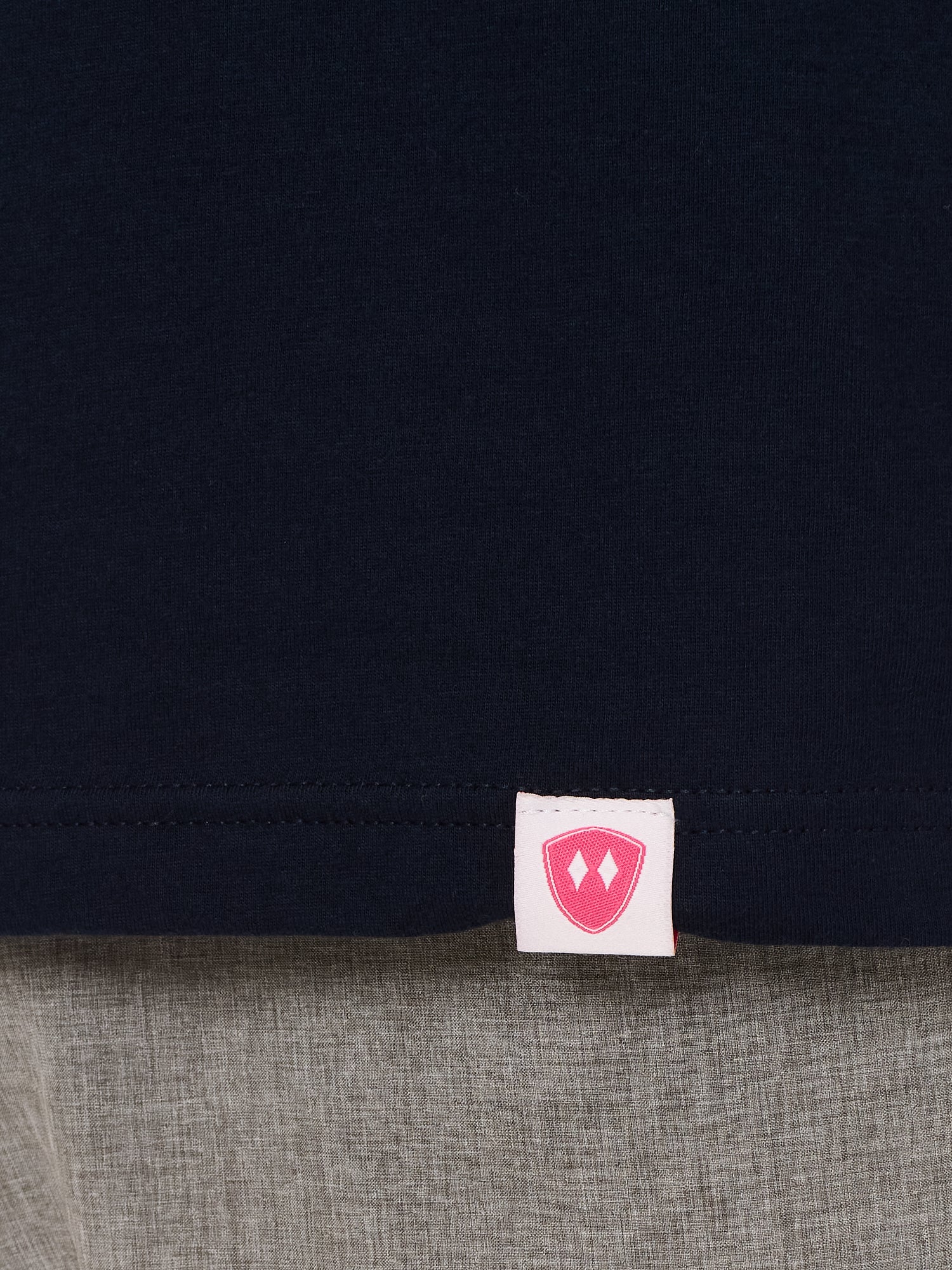 Close-up detail of a pink Double Diamond Shield woven hem label on a navy blue  shirt.