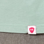 Close-up detail of a pink Double Diamond Shield woven hem label on a dusty blue short sleeve shirt.