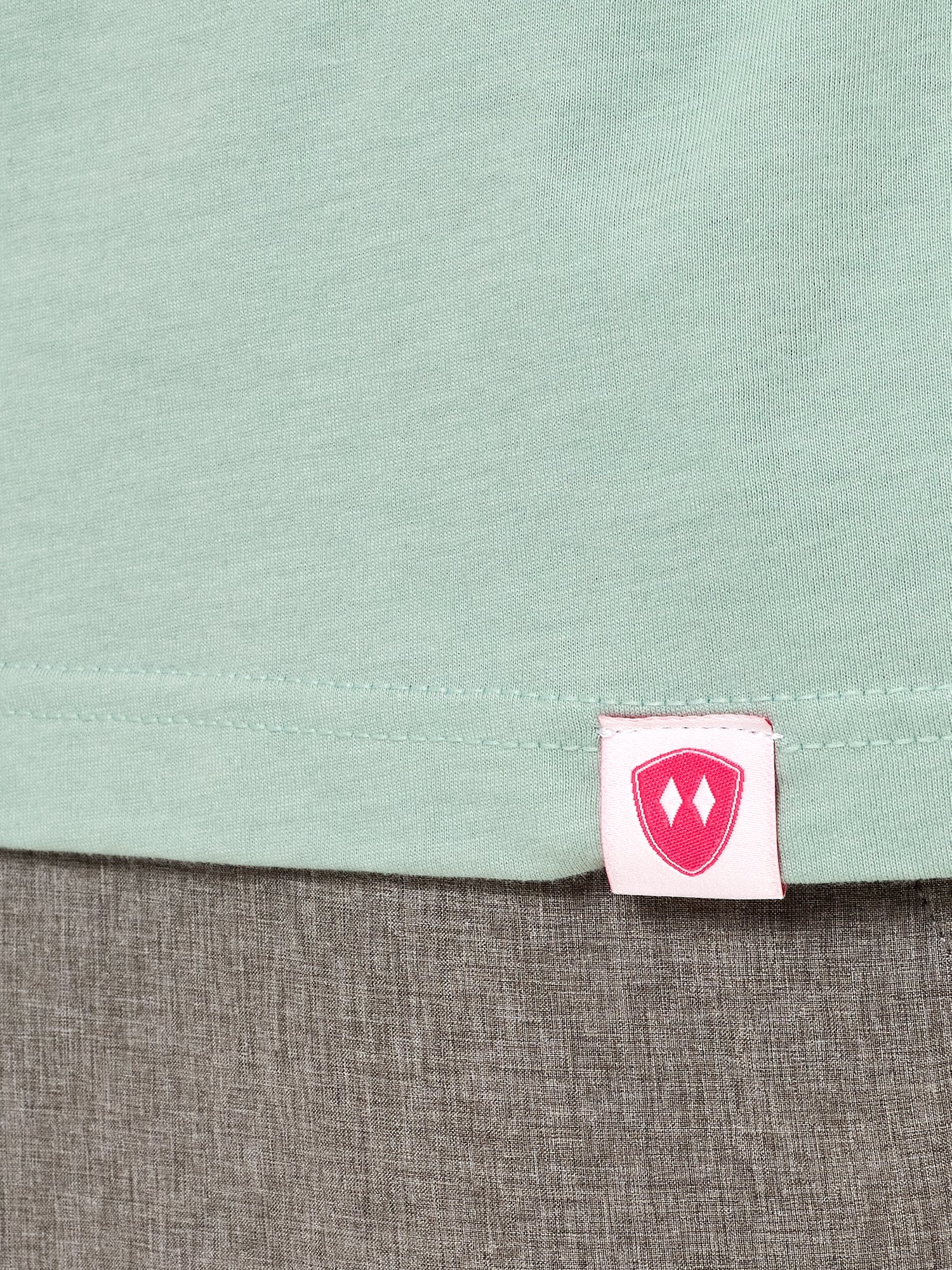 Close-up detail of a pink Double Diamond Shield woven hem label on a dusty blue short sleeve shirt.