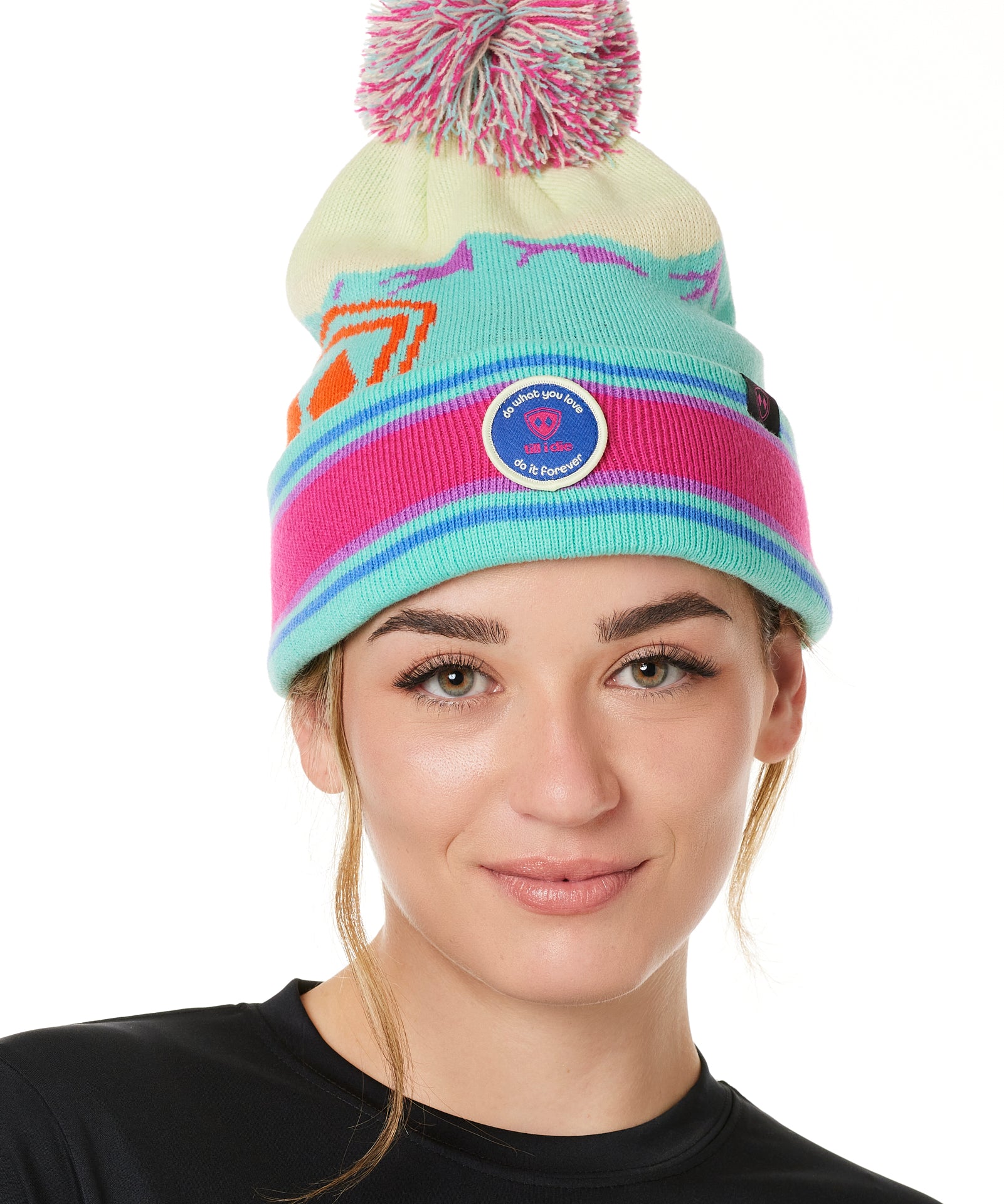 A WOMAN wearing a multicolored beanie with a pink pom-pom on top, featuring a TILL I DIE brand patch on the fold THAT SAYS "DO WHAT YOU LOVE. DO IT FOREVER."