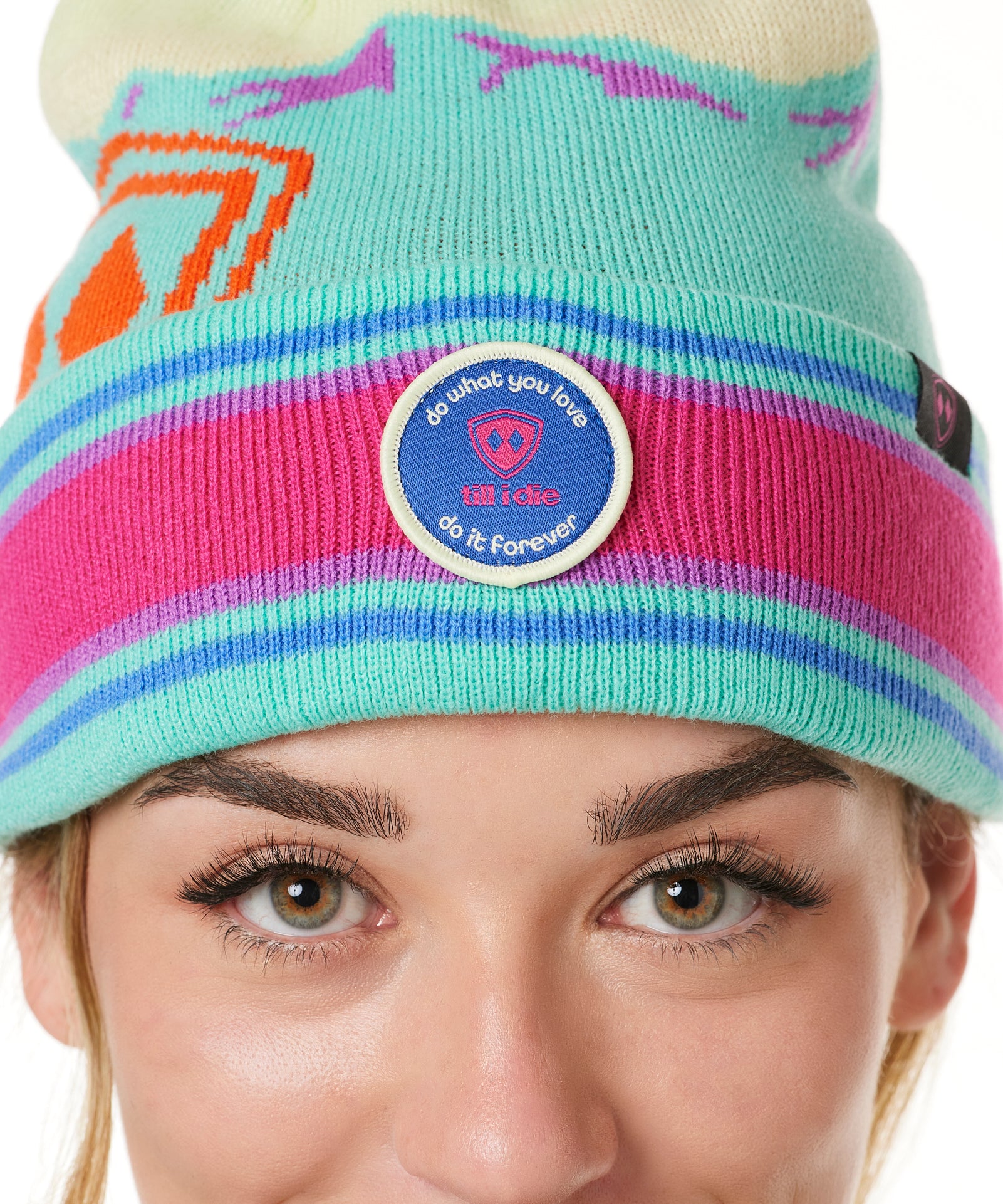 DETAIL A WOMAN wearing a multicolored beanie with a pink pom-pom on top, featuring a TILL I DIE brand patch on the fold THAT SAYS "DO WHAT YOU LOVE. DO IT FOREVER."