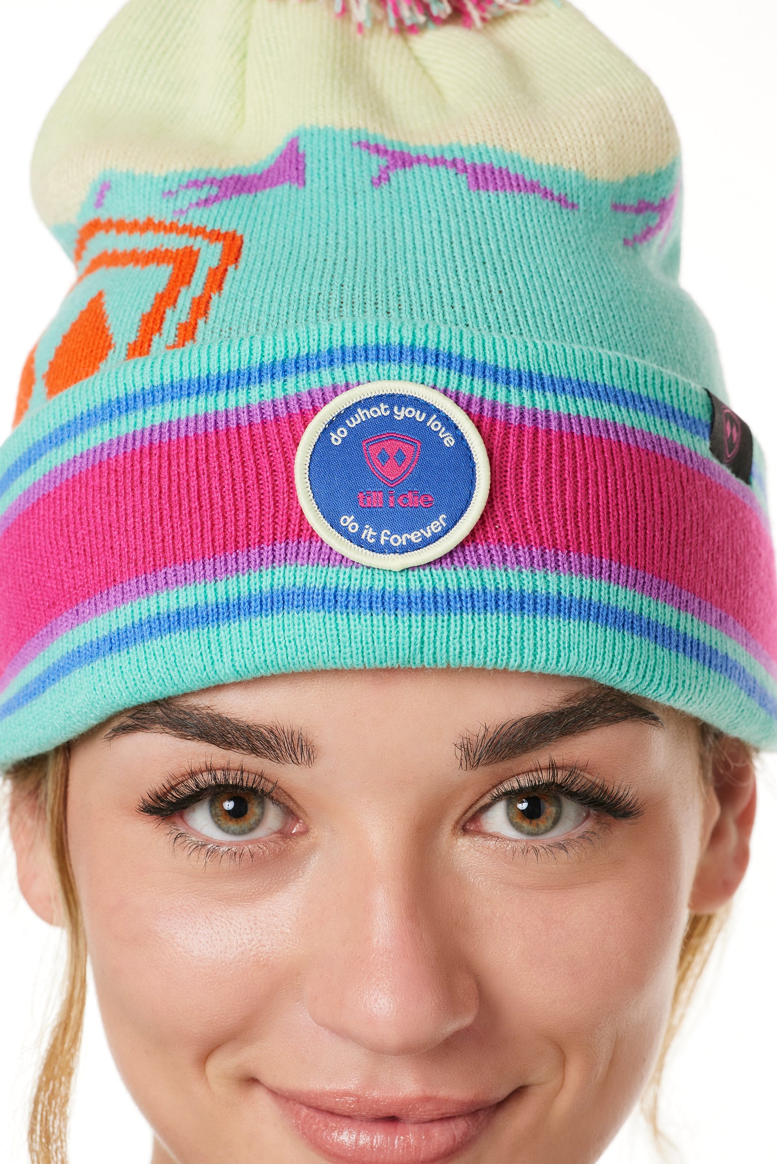 DETAIL A WOMAN wearing a multicolored beanie with a pink pom-pom on top, featuring a TILL I DIE brand patch on the fold THAT SAYS "DO WHAT YOU LOVE. DO IT FOREVER."