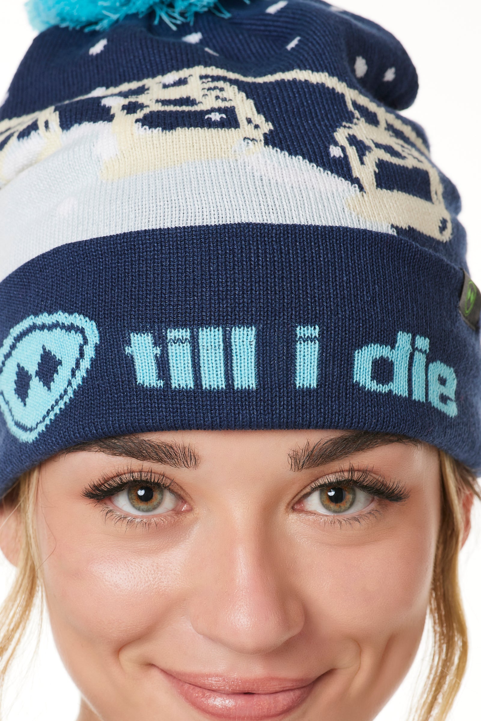 DETAIL OF A FEMALE MODEL WEARING A NAVY AND WHITE HAT WITH A BLUE POM POM AND CHAIRLIFT GRAPHIC WITH THE WORD "TILL I DIE" ON THE FOLD