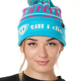 FEMALE MODEL WEARING A TEAL AND PINK HAT WITH A POM POM AND THE WORDS "APRES SKI"