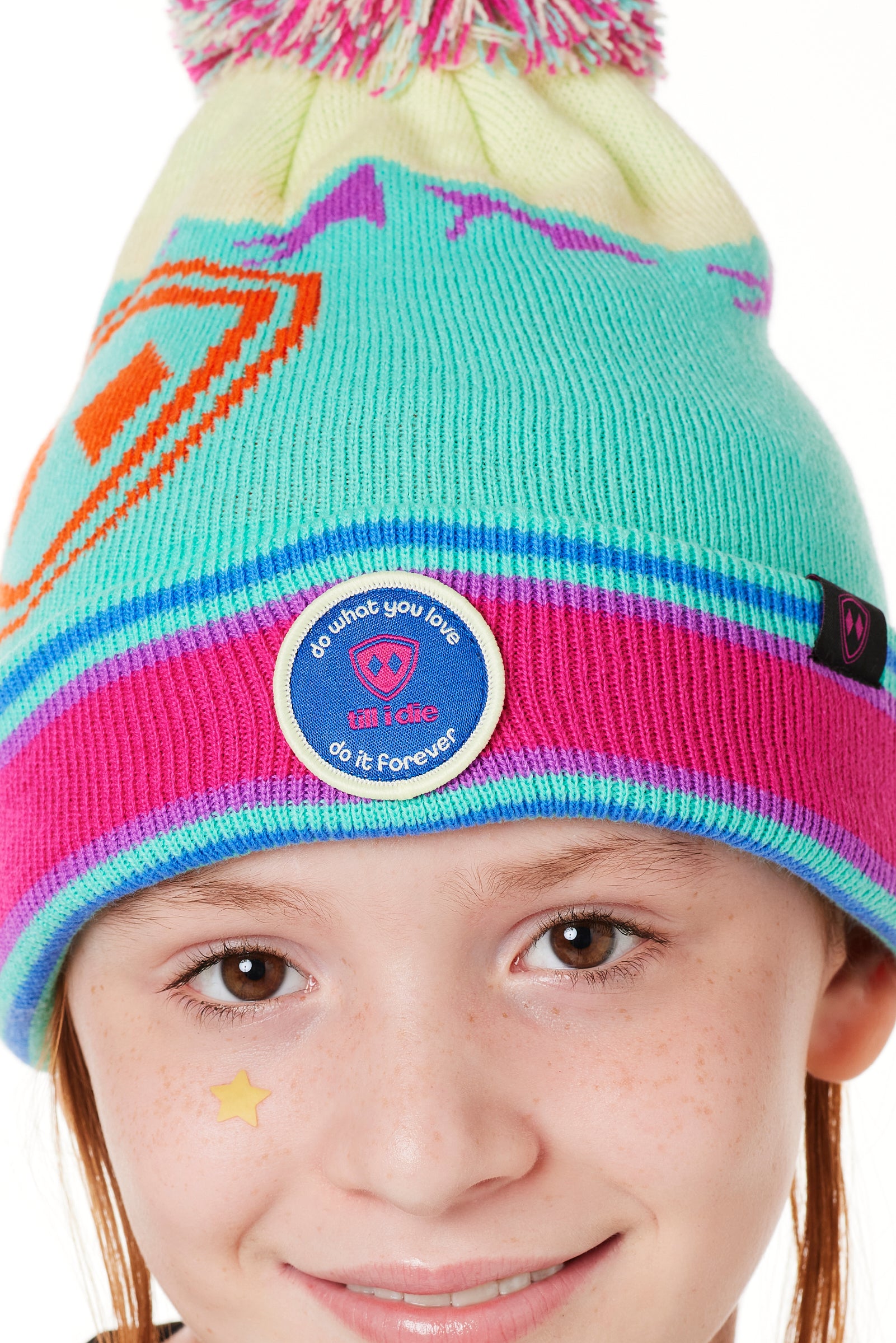 DETAIL OF A YOUTH wearing a multicolored beanie with a pink pom-pom on top, featuring a TILL I DIE brand patch on the fold THAT SAYS "DO WHAT YOU LOVE. DO IT FOREVER."