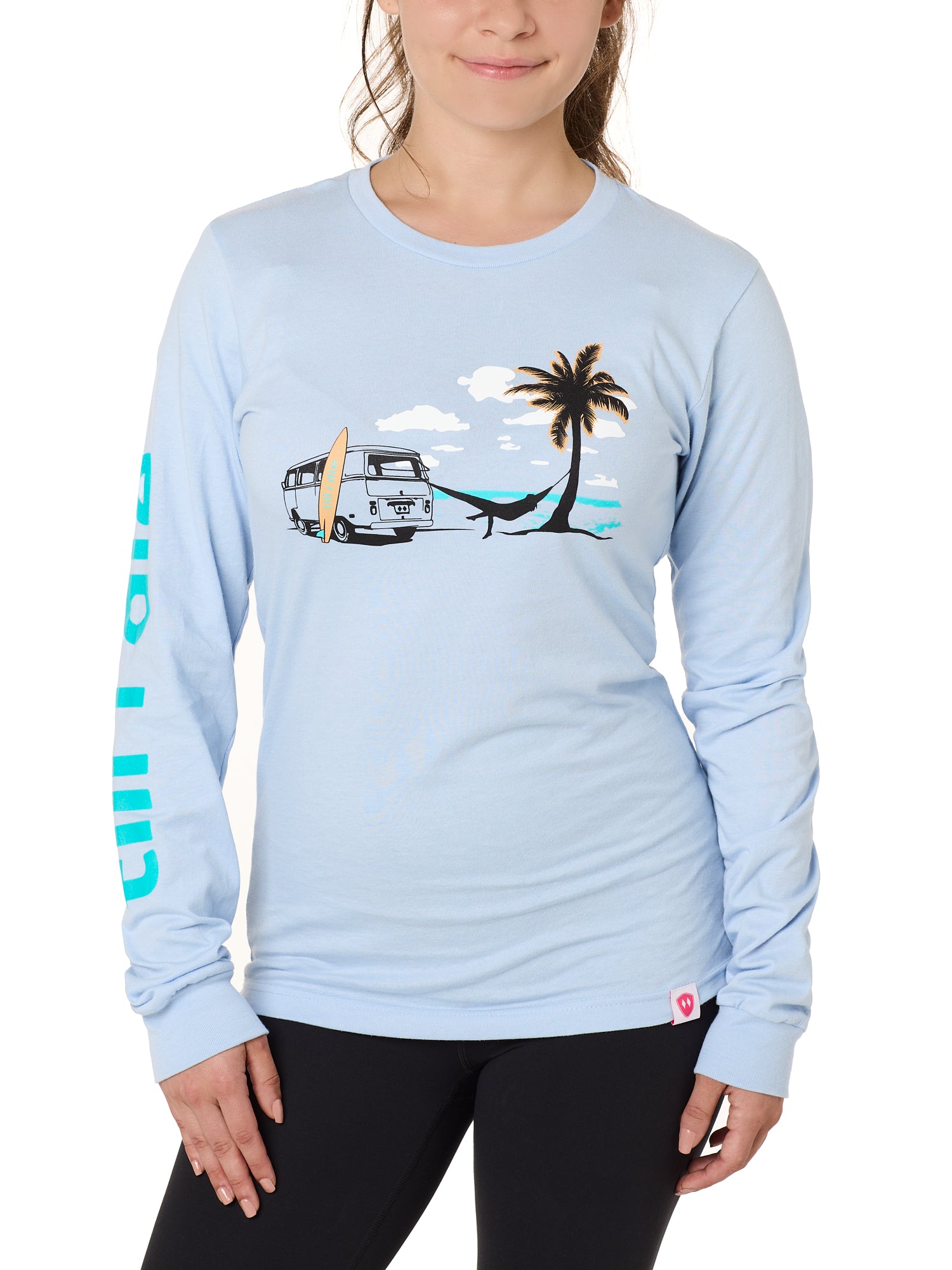 Front view of a female model wearing the Hammock long sleeve tee in light blue, displayed on a white background. The graphic depicts a vintage VW bus parked on a beach, with a surfboard leaning against it and a silhouetted figure relaxing in a hammock strung between the van and a palm tree.