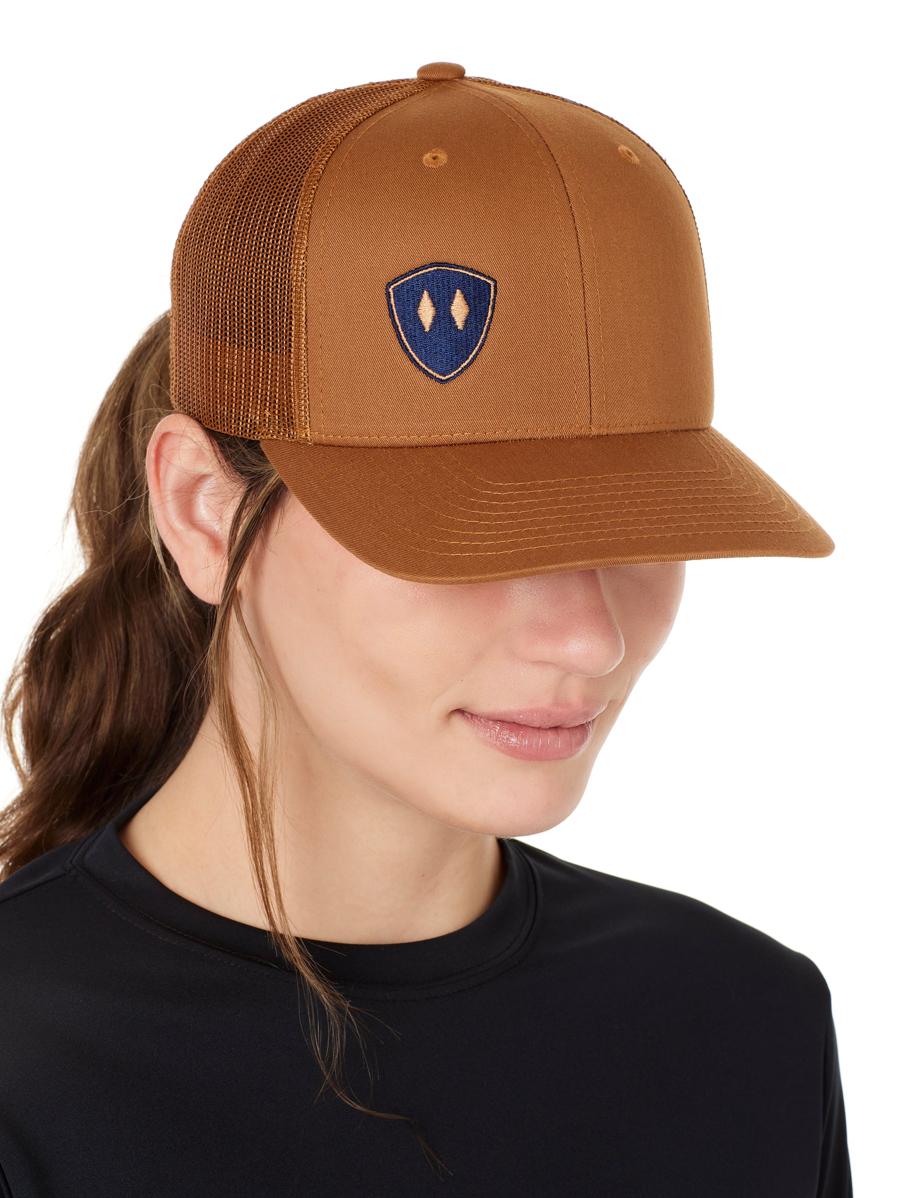 Front view of a female wearing a caramel classic trucker hat with a navy blue embroidered double diamond shield on the bottom right panel