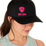 Front view of a female model wearing a black classic trucker hat, featuring a hot pink embroidered Till I Die logo on the lower right front panel.