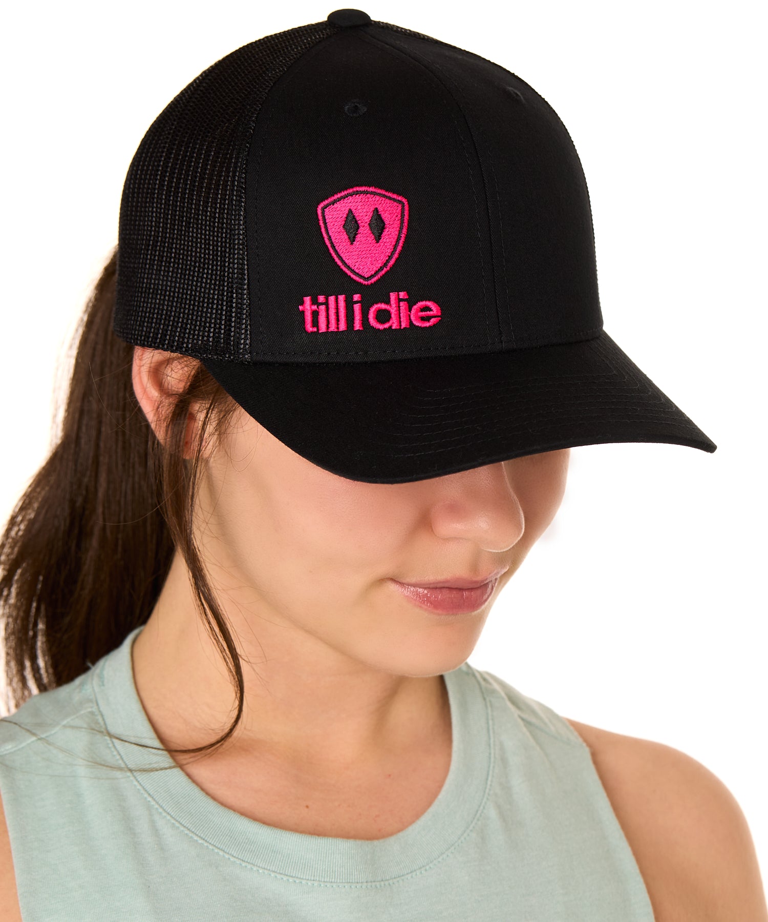 Front view of a female model wearing a black classic trucker hat, featuring a hot pink embroidered Till I Die logo on the lower right front panel.