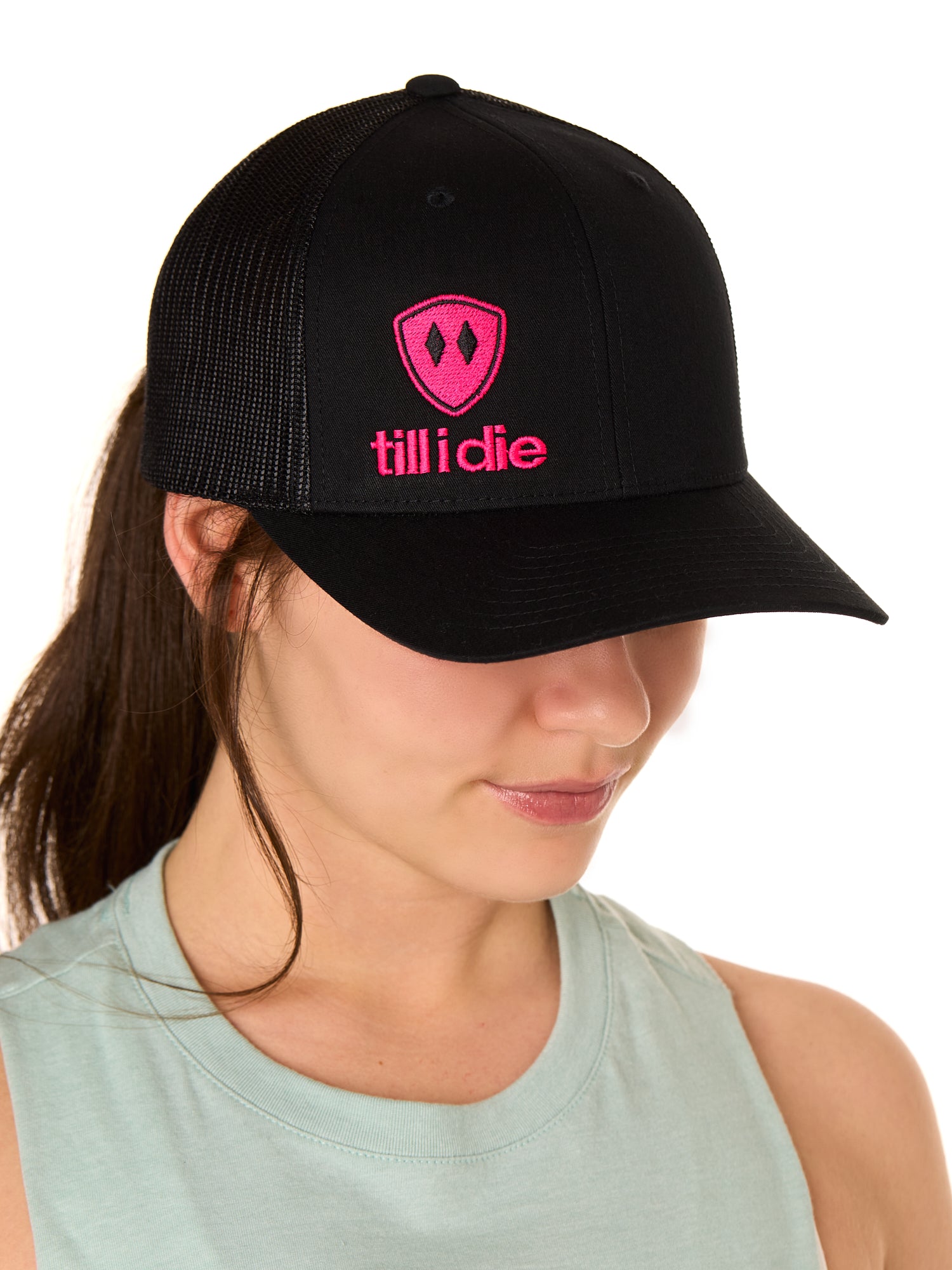 Front view of a female model wearing a black classic trucker hat, featuring a hot pink embroidered Till I Die logo on the lower right front panel.