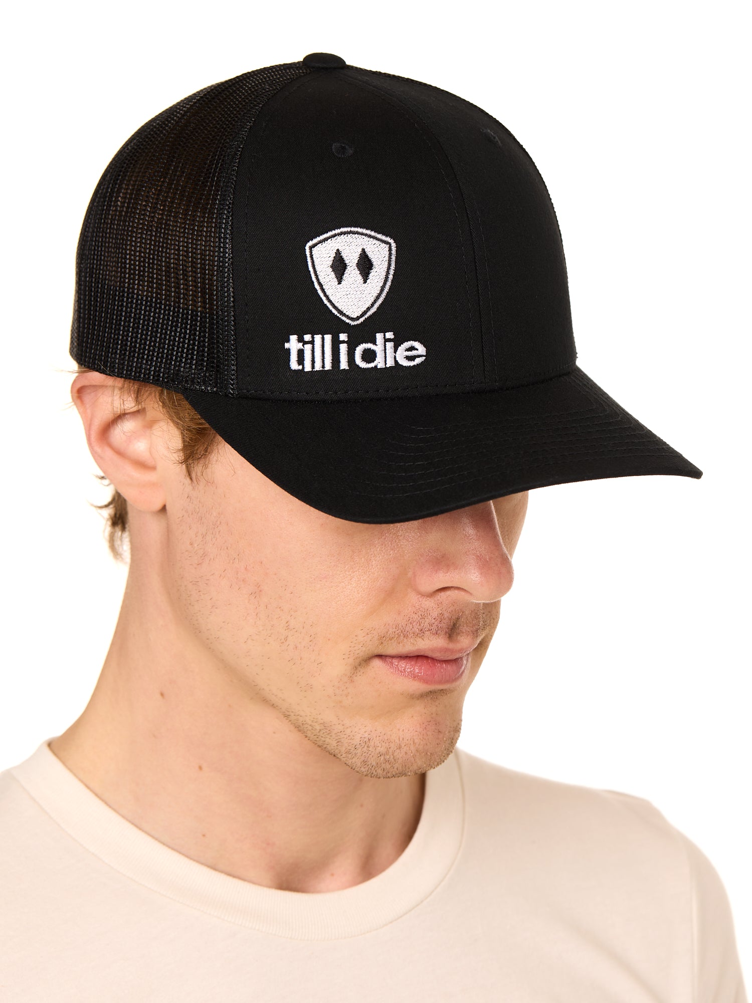 Front view of a male model wearing a black classic trucker hat, featuring a white embroidered Till I Die logo on the lower right front panel.